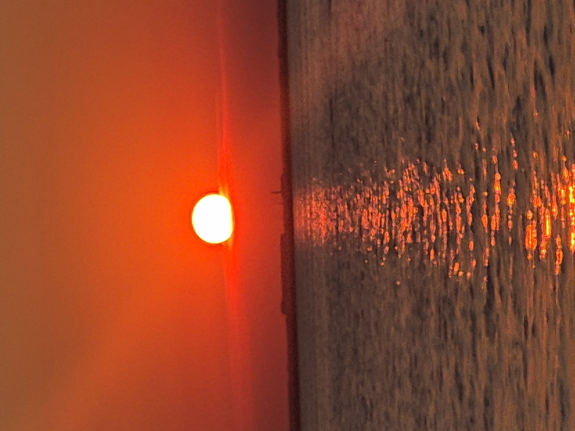 A sunset over a body of water. The sun is a bright orange orb partially obscured by a thin band of clouds, casting a fiery reflection on the water's surface. The sky is a gradient of orange and red, fading into a darker hue towards the top of the frame. A distant, low-lying landmass with a small structure is visible on the horizon. The water is choppy, with ripples distorting the reflection of the sun. The scene is devoid of any human presence. The location is the coast of the city of Kokshetau, Kazakhstan, situated on the shores of Lake Borovoye.
