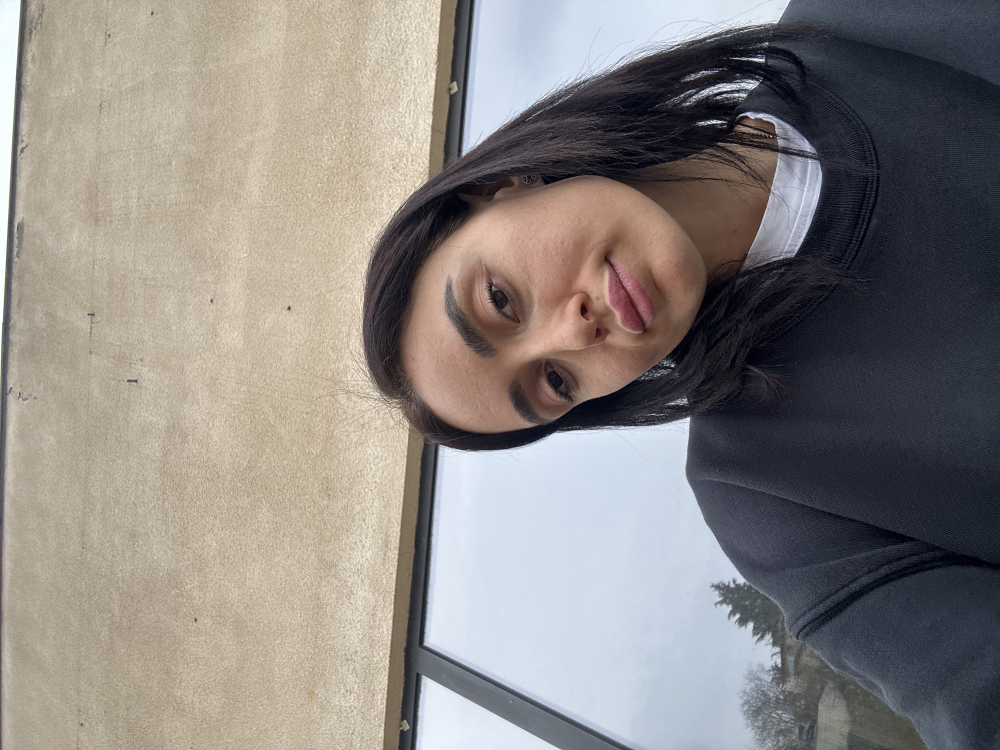 An upward-angled close-up selfie shows a female subject, approximately 20-30 years old, with dark brown hair and eyes, and a neutral expression. She wears a dark crewneck sweatshirt over a white t-shirt, with a dark stud earring visible in her right earlobe. The background consists of a light brown, textured building facade above a large, black-framed window. The window reflects an overcast sky and bare deciduous trees, indicating a cooler season. This scene is located in the city of Tbilisi, Georgia.
