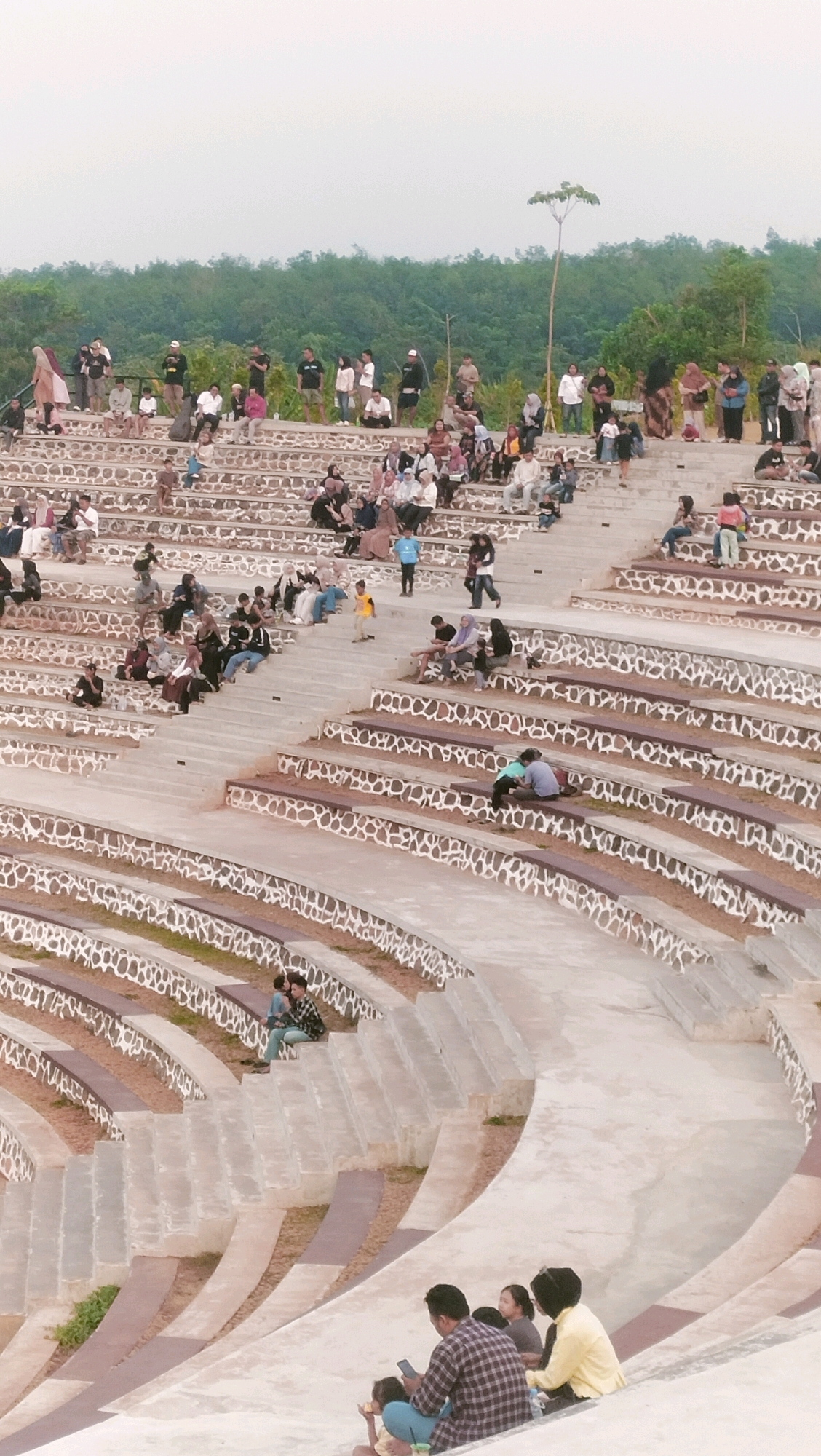 The image captures a tiered amphitheater setting with a crowd of people seated and standing. The amphitheater appears to be constructed with concrete seating areas and stone accents along the edges of each tier. Green trees and foliage form a backdrop behind the upper tiers of seating. The sky is overcast with a light gray hue.
In the foreground, several individuals are seated on the lower tiers. A man in a plaid shirt is looking at his phone, with two other people seated next to him. Further up and to the left, a couple is seated together, with one person leaning against the other.
As the view moves upwards, more people are visible on various tiers. Some are sitting, while others are standing and socializing. There are individuals and small groups scattered throughout the seating area. The crowd appears to be observing something or simply enjoying the ambiance of the location.
The overall scene suggests a public gathering or event taking place in an outdoor amphitheater in Ketanggan, Indonesia. The atmosphere seems relaxed and casual, with people interacting with each other and enjoying their surroundings.
