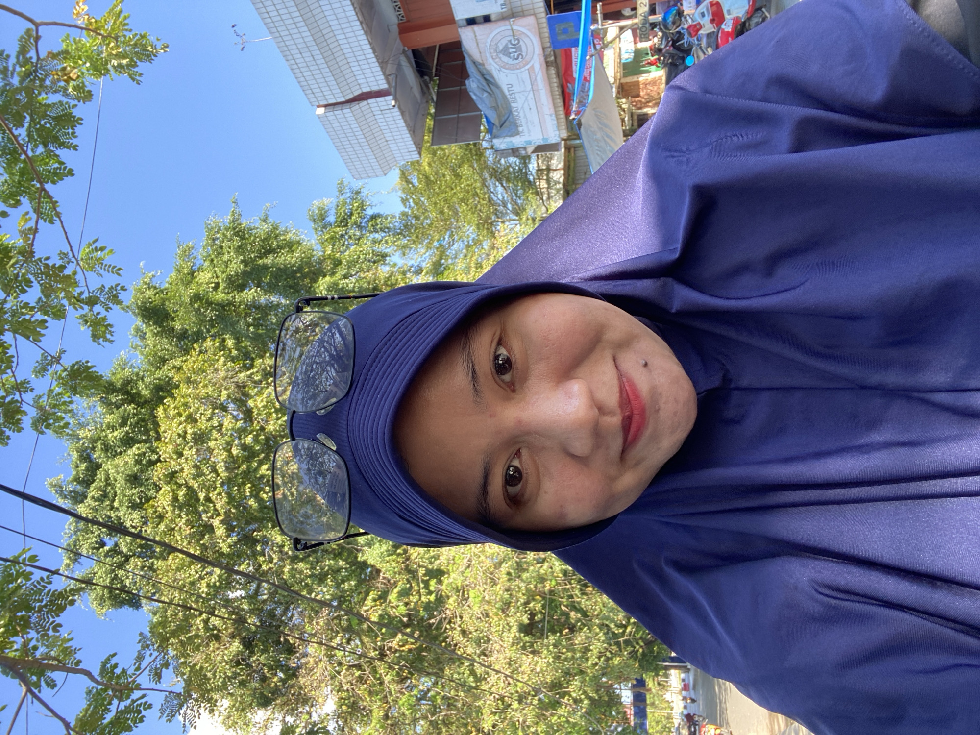 The image shows a selfie of a woman wearing a dark blue hijab and glasses. She is outdoors with a bright blue sky and lush green trees in the background. A building with a white tiled facade and red accents is visible to the right, along with signs that appear to be in Indonesian. The overall lighting suggests it is daytime. The presence of the building and the visible signage indicate an urban or semi-urban setting in Martapura, Indonesia.