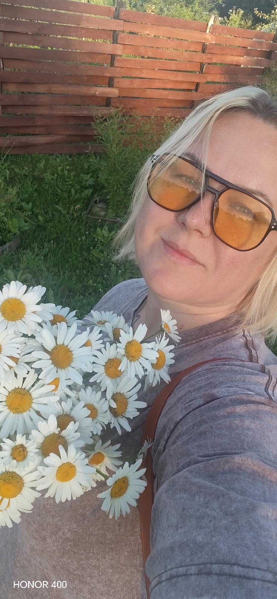 A woman with short blonde hair and sunglasses with orange lenses is holding a bouquet of daisies. She is wearing a gray t-shirt. The background shows a wooden fence and green grass, suggesting an outdoor setting, possibly a garden or rural area. The lighting indicates it might be late afternoon or early evening. A brown strap is visible across her shoulder. The image contains the text "HONOR 400" in the bottom left corner. The overall mood appears relaxed and pleasant.