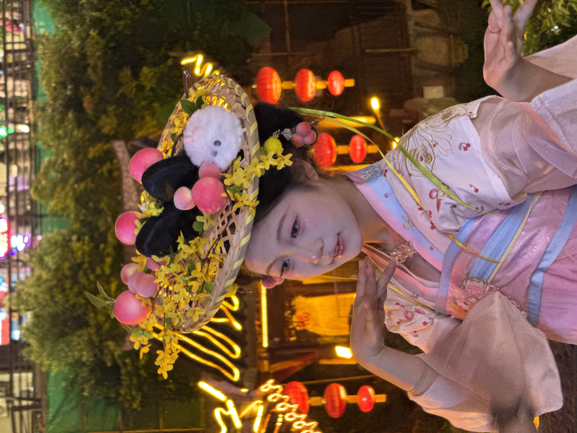 A young East Asian woman, appearing to be in her late teens or early twenties, is depicted in the foreground against a softly lit, outdoor nighttime setting. She is wearing a traditional Chinese-style outfit in shades of pink, blue, and white, featuring wide sleeves and intricate embroidery. Her hair is styled elaborately, adorned with a woven basket-like headdress that is decorated with pink faux peaches, yellow flowers, black bows, and a small white fluffy creature resembling a bunny. She has makeup on, including glitter on her eyelids and cheeks, and her expression is gentle and somewhat wistful. Her right hand is raised near her face in a graceful pose, while her left hand is extended outward.
The background suggests an urban environment at night, likely in Yuzhong District, China, given the presence of traditional red lanterns hanging in clusters, and illuminated neon signs or decorative lights forming abstract shapes. Lush green foliage is visible behind the woman. The overall atmosphere is festive and decorative, possibly related to a cultural event or holiday.