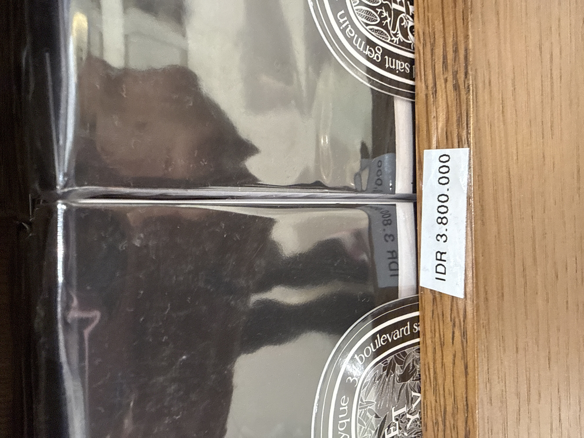 The image shows two reflective rectangular boxes side-by-side on a wooden shelf. A white price tag displaying "IDR 3.800.000" is adhered to the shelf in front of the boxes. The boxes have metallic surfaces that reflect a dark environment, with some subtle hints of what might be the interior of a shop. On the lower part of the left box, a circular label with text and an illustration is partially visible, reading "boulevard s". On the right box, a similar circular label is visible with the text "saint germain". The overall scene suggests a retail setting, likely a boutique or department store, and the currency indicates the location is in Indonesia. The image does not provide specific details about the city of Kuta.