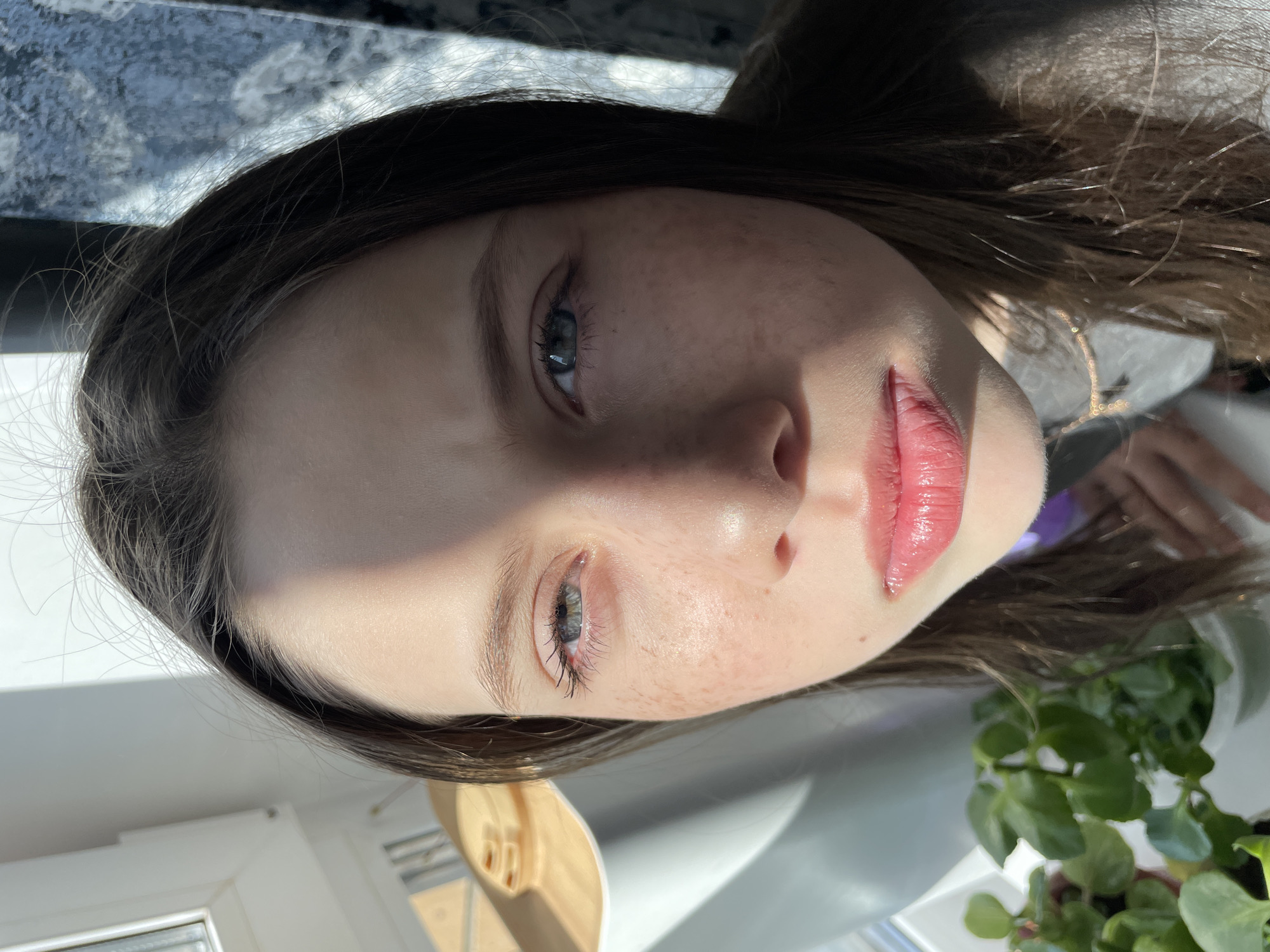 A young woman with fair skin and freckles is the primary subject of the image. She has brown hair and light-colored eyes. Her lips appear to have some kind of gloss or lip color on them. The image is taken from a low angle, looking up at her face. Part of a window with a white frame is visible, along with a potted green plant. Based solely on the provided image, it's not possible to confirm if the location is in Izhevsk, Russia. There is no indication of an action or interactions in the image.