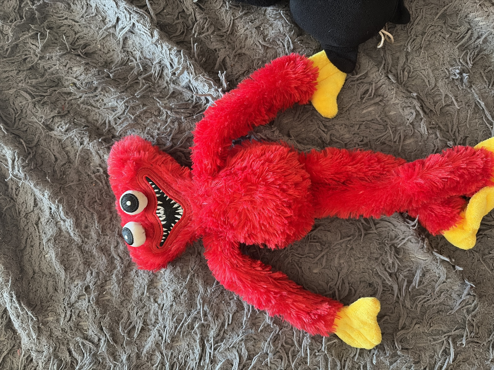 A red, fuzzy toy with yellow feet is lying on a grey, shaggy rug. The toy has two large, white eyes with black pupils, and a wide, toothy grin with black and white jagged teeth. Its body is elongated, and its limbs are spread out. In the background, a portion of a black plush toy is visible. The setting appears to be indoors, likely a bedroom or living space, given the presence of the rug and plush toys. There are no discernible cues about the time of day, weather, or specific emotions conveyed by the scene, other than the slightly menacing appearance of the red toy. No visible text is present in the image.
