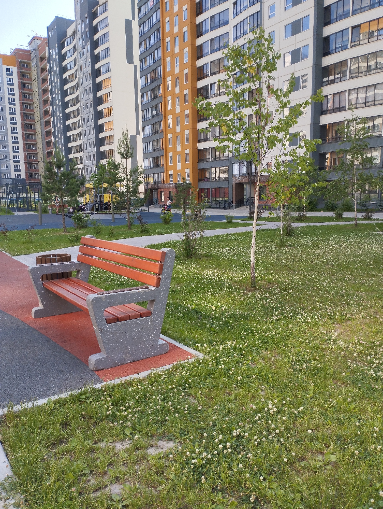 The image depicts a modern residential courtyard in Sadovyy, Russia, featuring a playground, a bench, and apartment buildings. In the foreground, a contemporary wooden and concrete bench is positioned on a paved area adjacent to a grassy lawn dotted with small white flowers. The bench is empty. Behind the bench, a playground with colorful structures is visible, and several children are present, engaged in play. Further in the background, a row of tall, multi-story apartment buildings with contemporary architecture dominates the skyline. The buildings are a mix of orange, gray, and white facades with numerous windows. Trees are scattered throughout the courtyard, some small and sapling-like, others more mature. The overall impression is of a well-maintained, family-friendly urban environment.