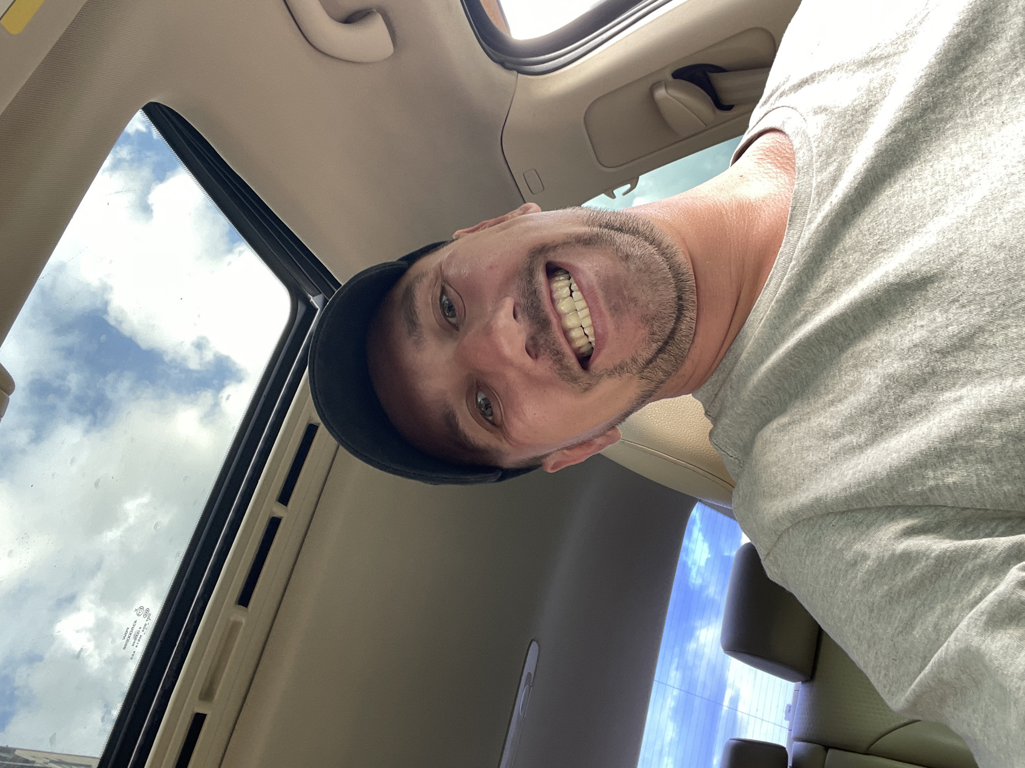 A man wearing a black baseball cap and a grey t-shirt is inside a car, looking at the camera. The car has a sunroof open, revealing a cloudy sky. The interior of the car appears to be beige. The man has a goatee and is smiling, showing his teeth. The image is taken from a low angle. The background shows the rear seats of the car and the sky through the rear window. There is no discernible location within the city of Kyiv, Ukraine.