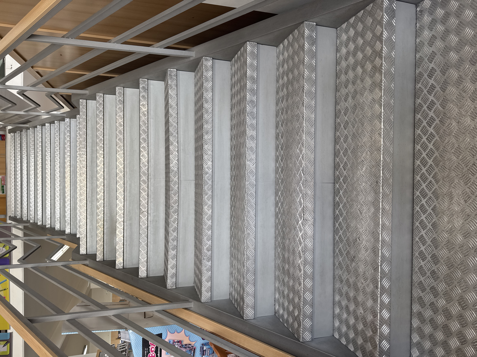 The image shows a metal staircase with diamond plate treads, located in Agios Athanasios Municipality, Cyprus. The staircase is part of an indoor or covered outdoor structure, evidenced by the presence of a wooden handrail and metal railing on the left side, and a wooden paneled wall on the right. The staircase is unoccupied and appears to be in good condition. The lighting suggests it could be daytime, with light reflecting off the metal surfaces. The background is slightly out of focus but suggests a commercial or public building setting, possibly a library or a school, with shelving and promotional materials visible on the far left.
