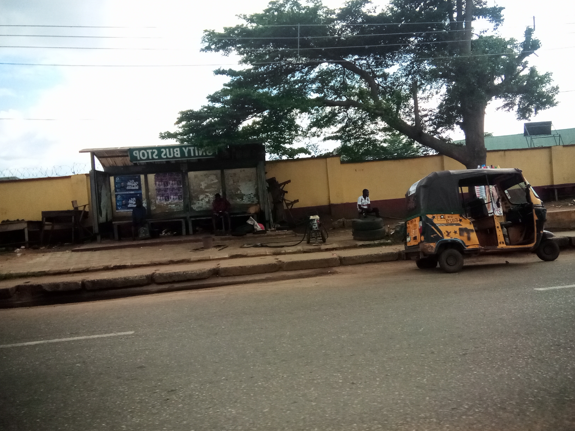 The image shows a street scene in Nibo, Nigeria, with a bus stop on the left and a yellow auto-rickshaw on the right. The bus stop has a covered shelter with benches and advertisements displayed on the walls. Several people are seated on the benches at the bus stop. Behind the bus stop, a yellow wall with barbed wire on top runs along the property line, and large trees with dense foliage arch over the scene. In front of the bus stop, a sidewalk with curbs separates the bus stop area from the road. On the right side of the image, the yellow auto-rickshaw is parked on the side of the road, its doors open and its canopy extended. Another person is seated on the curb to the left of the auto-rickshaw. The road in the foreground is asphalt with a white line marking its edge. The sky is overcast with some blue sky visible.