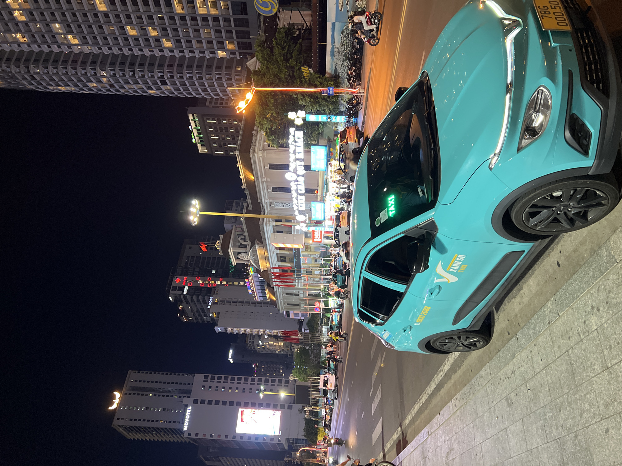 The image depicts a vibrant street scene at night in Nha Trang Ward, Vietnam. A turquoise Xanh SM taxi is prominently featured, parked on the right side of the frame. The cityscape is illuminated by streetlights and the glow of buildings, including the Novotel Nha Trang. The license plate of the taxi reads "78G 000.50." Traffic, primarily scooters, can be seen moving along the road. Various commercial establishments and signage line the street, adding to the urban atmosphere.