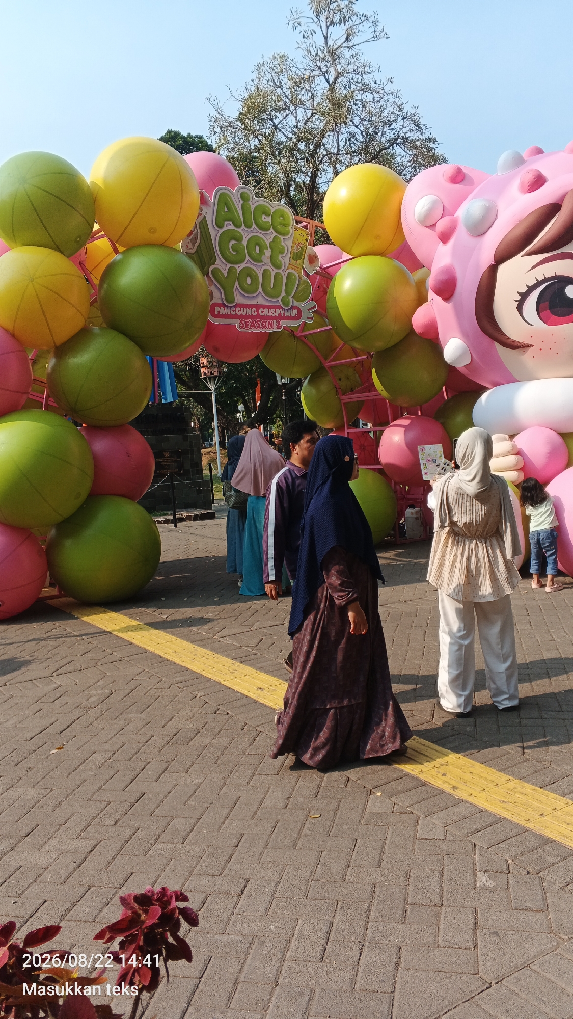 An outdoor, daytime scene depicts a festive archway constructed of large, colorful balloons in yellow, green, and pink. The arch is decorated with a sign that reads "Aice Got You! Panggung Crispy! Season 2." To the right, a large, pink cartoon character, possibly a mascot, is visible. Several people are present, including a woman in a dark blue hijab and a patterned dress, a man in a dark jacket, and another woman in a light-colored hijab and pants. A young child in a light green shirt and blue jeans is standing near the pink mascot. The ground is paved with interlocking bricks and features a yellow tactile paving strip. The background shows trees and a clear blue sky. The image appears to be a street or public space, possibly part of an event or festival. A timestamp in the lower left corner reads "2026/08/22 14:41" and "Masukkan teks".