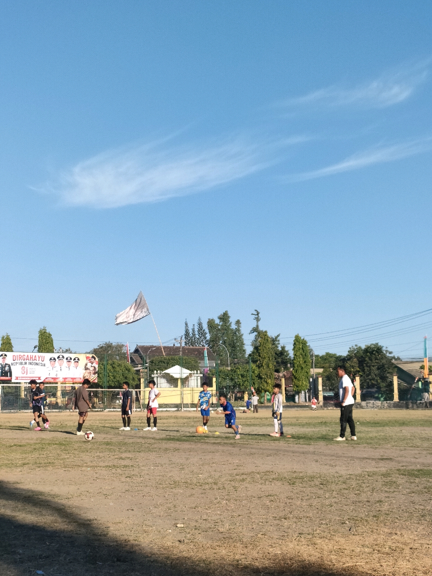This image depicts an outdoor soccer practice on a dry, dusty field in Kasihan, Indonesia, under a clear blue sky with wispy clouds. Several young boys are engaged in soccer drills, with one boy in a blue jersey actively running with the ball. Another boy in a brown shirt stands near a soccer ball, and a group of boys in various colored jerseys are positioned around the field. An adult male in a white t-shirt and black pants is standing on the right side of the frame, observing the practice. In the background, a large banner with Indonesian text and images of political figures is visible, along with trees, buildings, and a fence. A flag is also flying from a pole in the background. The overall scene suggests a local community event or sports activity taking place in the afternoon.