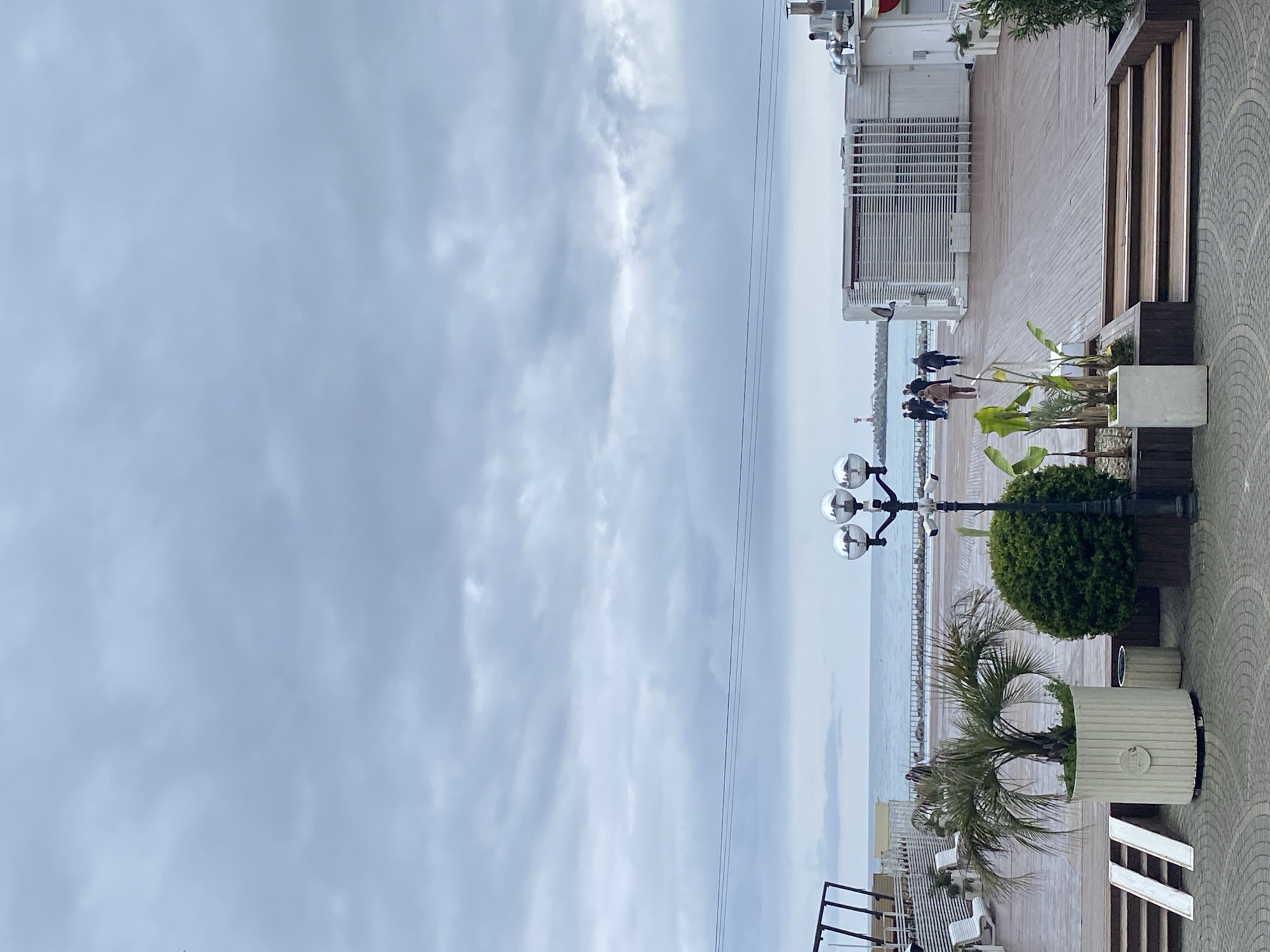 The image depicts a boardwalk in Sochi, Russia, under a cloudy sky. Several people are walking along the boardwalk near a breakwater. A distinctive lamp post with four globe lights and several large planters containing trees and other plants are visible. The boardwalk is made of light-colored wood. A white building with slatted windows is to the right of the frame. Benches are along the boardwalk.