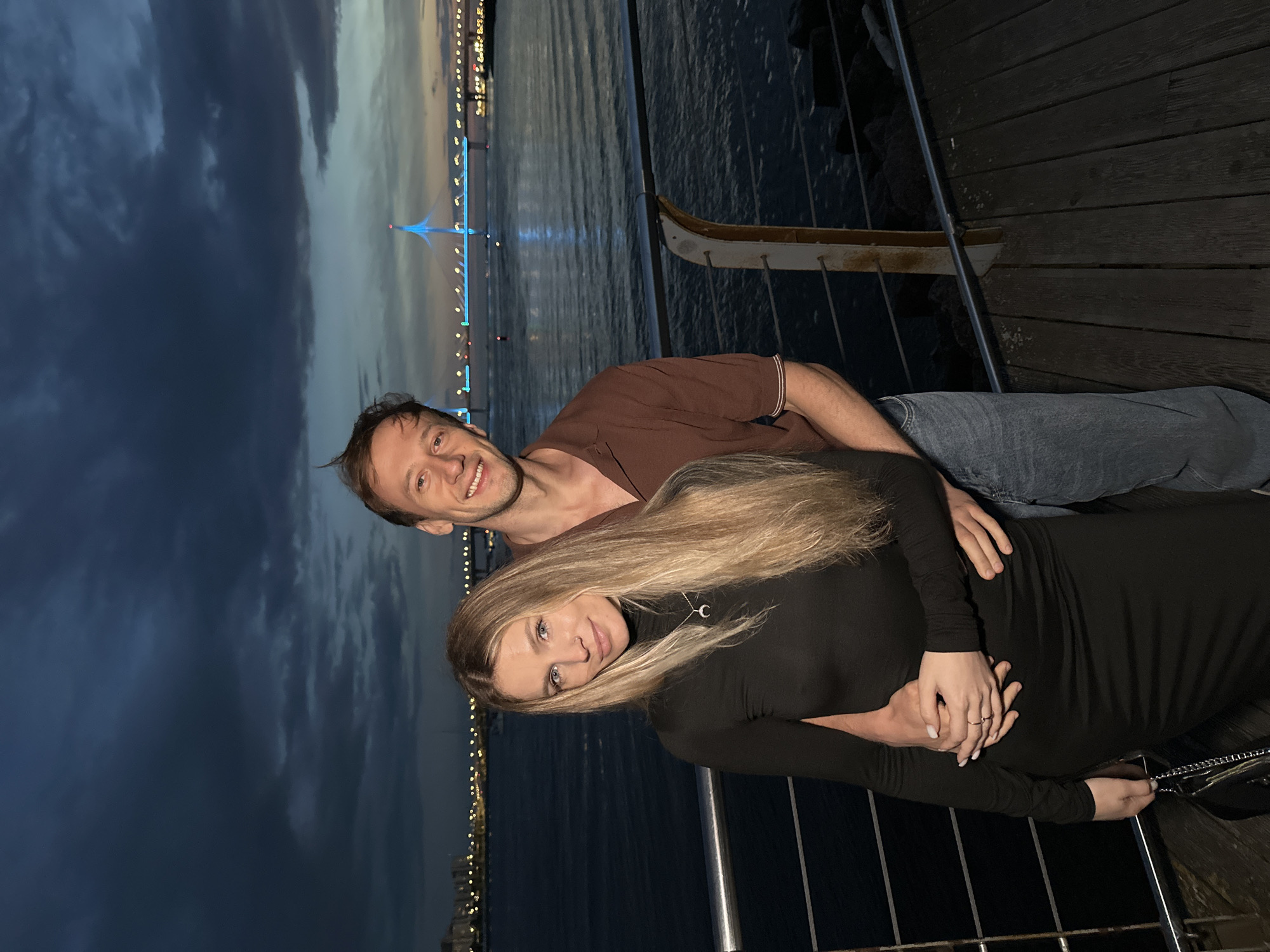 A man and a woman are standing on a wooden pier at dusk. The man, on the right, is wearing a brown polo shirt and blue jeans. He has light brown hair and is smiling at the camera. The woman, on the left, is wearing a long-sleeved black dress. She has long blonde hair and is looking at the camera with a slight smile. The man has his arm around the woman. In the background, there is a body of water, with a bridge illuminated by blue lights and city lights visible on the far shore. The sky is dark with some clouds. The location is the embankment of the Gulf of Finland in Saint Petersburg, Russia, possibly near the Western High-Speed Diameter bridge.