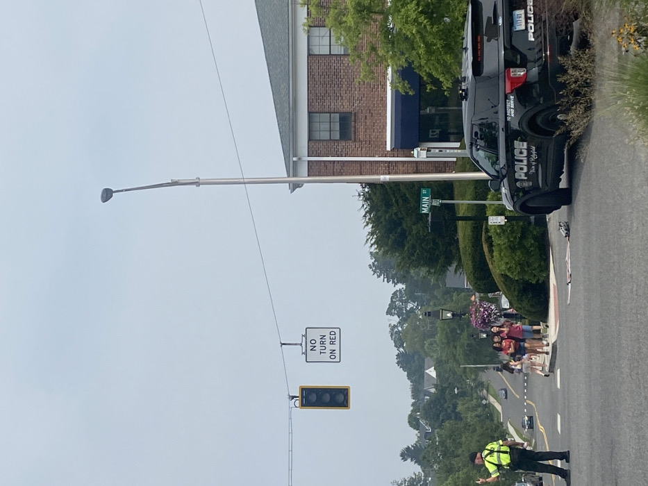 The image depicts a daytime street scene on Main Street in Ridgefield, United States, under an overcast sky. In the foreground-left, a male police officer, wearing a lime green reflective vest over a dark uniform and a dark cap, is standing with his right arm raised, signaling. A black Ford Explorer police SUV, marked "POLICE Town of Ridgefield" and bearing Connecticut license plate "117 RT," is parked on the right side of the roadway, facing away from the camera. Directly in front of the police vehicle on the pavement, a flattened cardboard box-like object and a grey cylindrical object are visible.
In the mid-ground, a group of four young female pedestrians are walking on the sidewalk from the background towards the viewer's left. Overhead, a traffic light displaying a yellow signal hangs alongside a "NO TURN ON RED" sign. A green street sign reading "MAIN ST" is visible on a pole on the right side of the road. A two-story brick building with white trim and a blue awning is located on the right. The environment includes mature trees, manicured bushes, and decorative street lamps, one featuring hanging baskets with purple flowers. Other residential structures are visible in the background, situated on a rising slope.