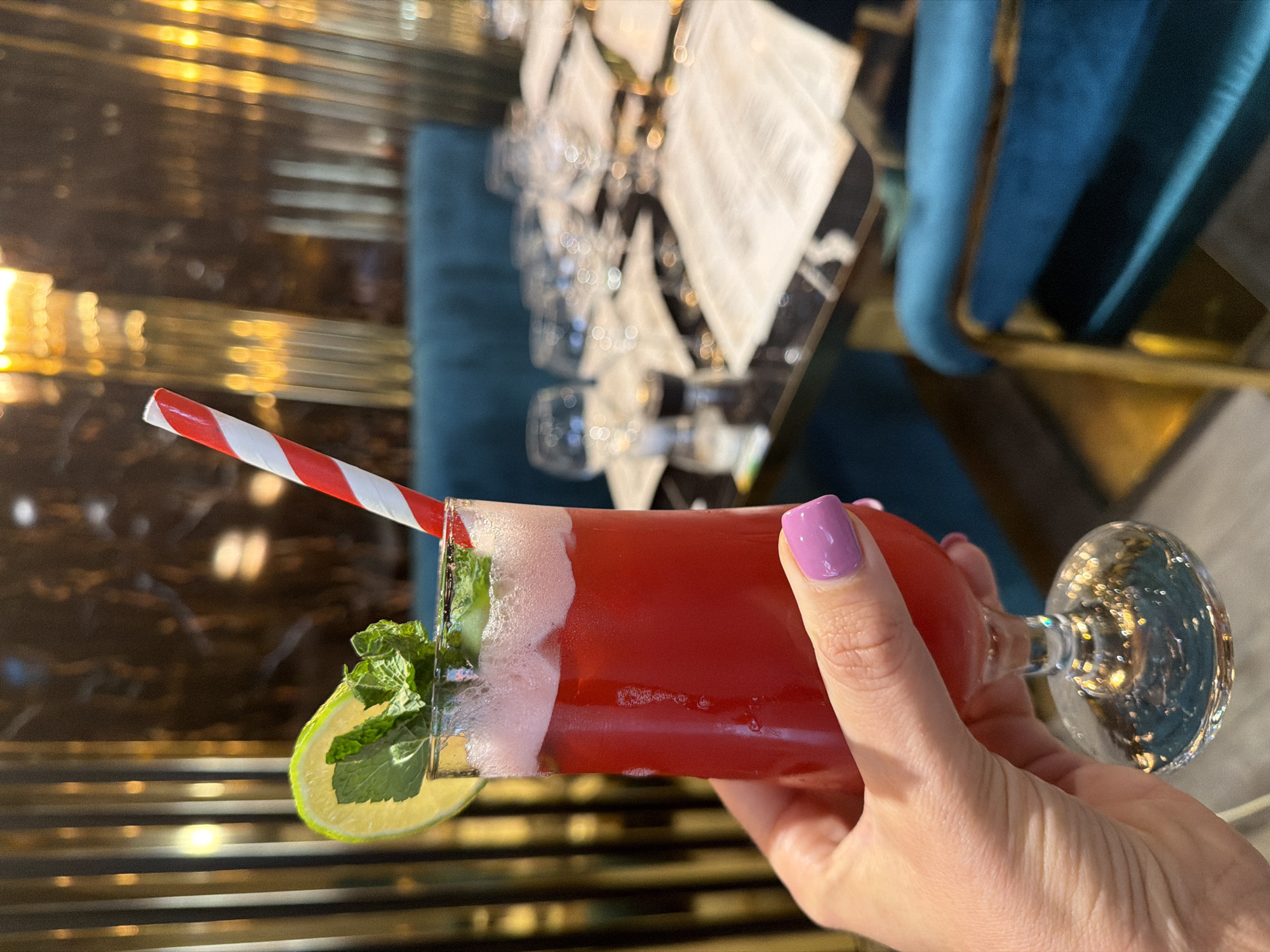 A hand holds a red cocktail in a clear glass garnished with mint and a lime slice, along with a red and white striped straw. The cocktail has a frothy white top. In the blurred background, there are dining tables with glasses and napkins and blue upholstered chairs. A mirrored surface with horizontal gold stripes is also visible. The location information "Mole Valley, United Kingdom" appears extraneous, as the image does not contain sufficient locational detail to determine a specific place.