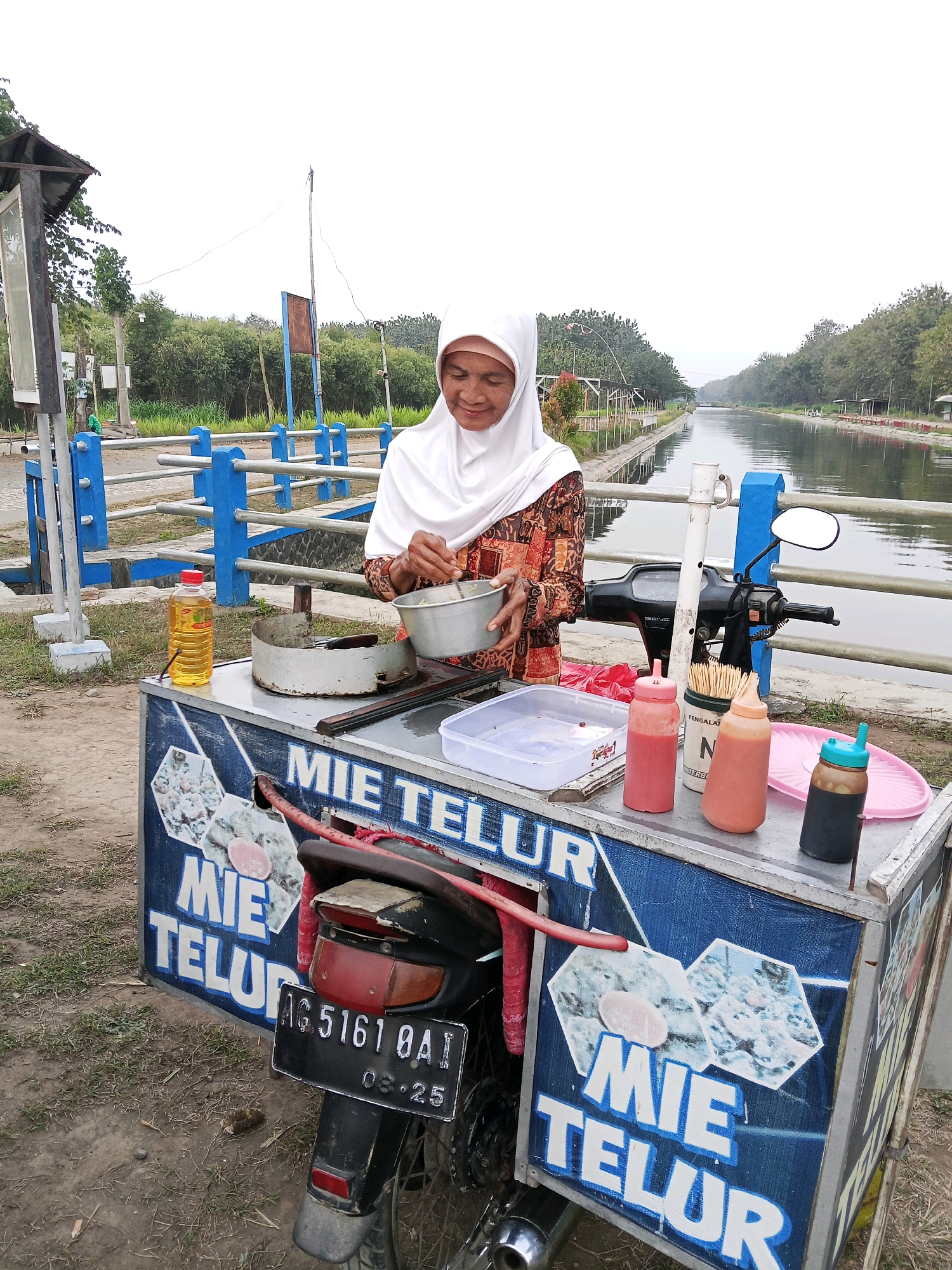 A woman wearing a white hijab and a patterned dress stands behind a food cart selling noodles in Darungan, Indonesia. The cart, decorated with blue panels and images of noodles, is attached to a motorcycle. On the cart are several bottles of sauce, a container of toothpicks, and a bottle of cooking oil. The woman is in the process of preparing food, holding a metal bowl. In the background, a canal runs through a tree-lined area. The sky is overcast.