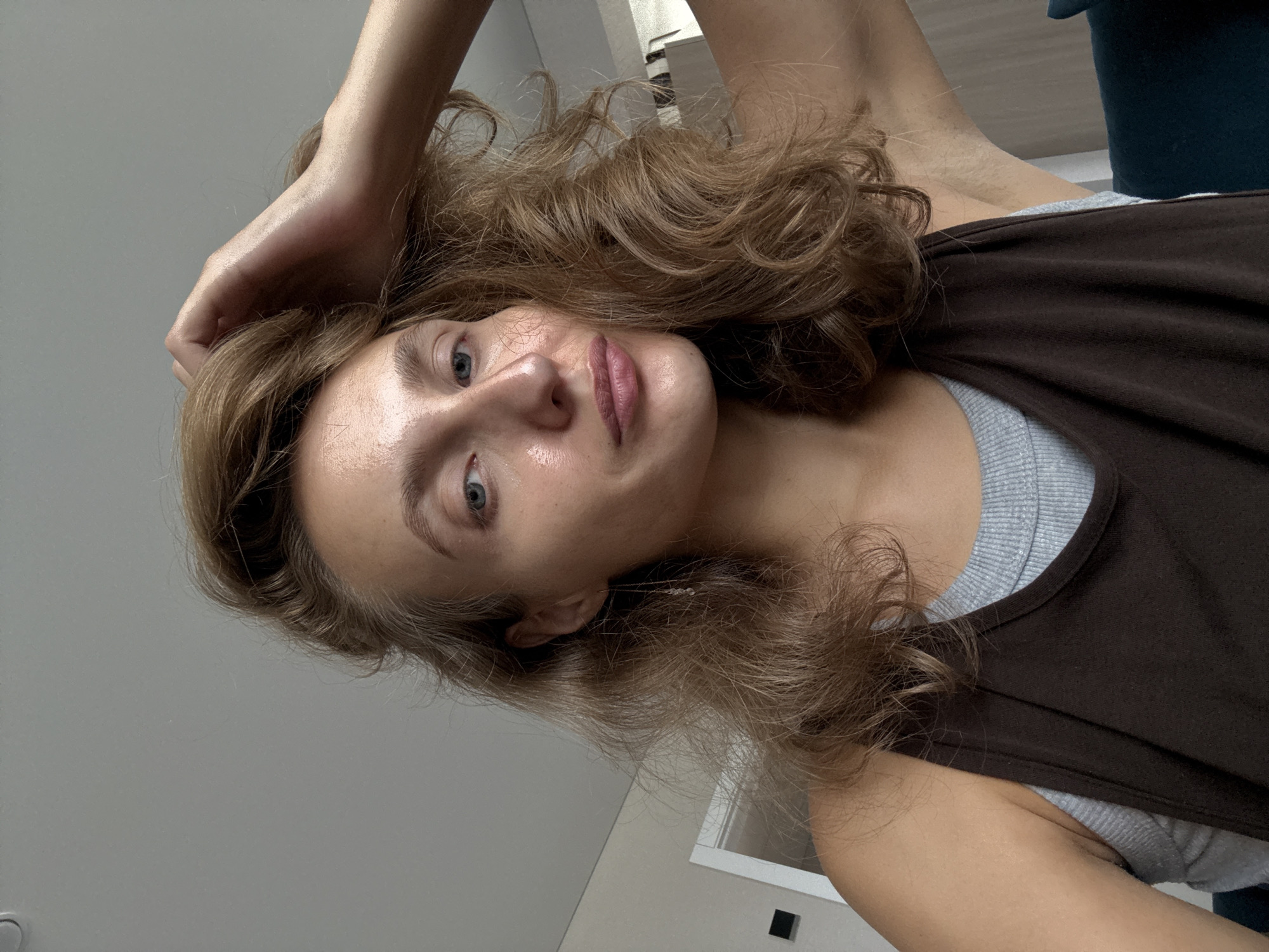 A woman with light brown hair and blue eyes is shown in a close-up selfie. She is wearing a dark brown tank top over a grey t-shirt. Her right hand is raised, touching her hair, and her left arm is bent. The background appears to be an indoor setting with a light grey wall and a ceiling. There is a door frame and a dark blue piece of furniture visible in the background. The lighting suggests an indoor environment. No specific location in the city of Подшивалово, Russia can be determined from the image.