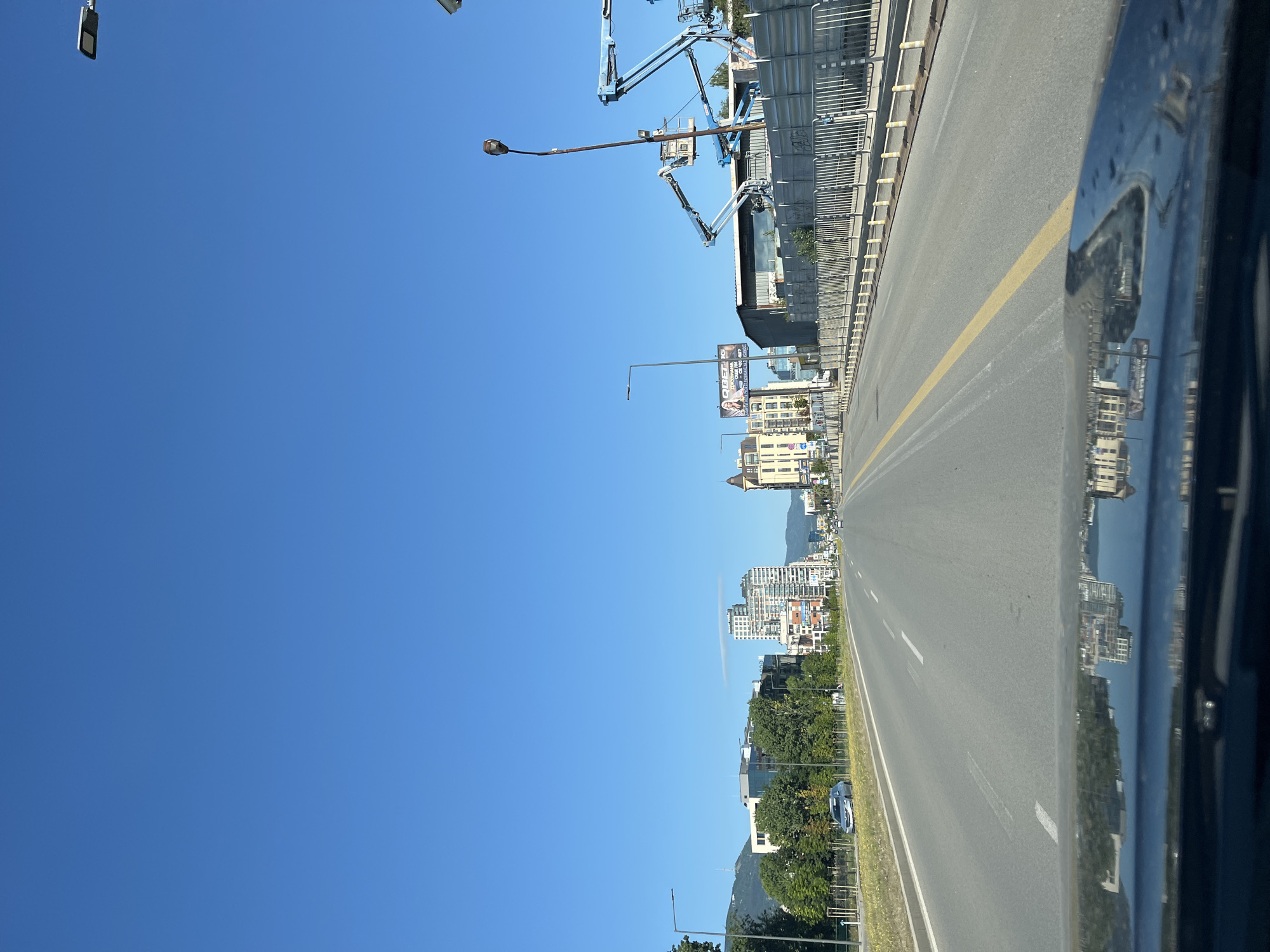 A frontal view from inside a vehicle shows a wide, multi-lane asphalt road in Sofia, Bulgaria, under a clear blue sky. The road stretches into the distance, bordered by a paved shoulder and a metal fence with yellow bollards on the right. On the left, trees and some low buildings are visible, with taller buildings and mountains in the background. The reflection of buildings and the sky can be seen on the vehicle's hood in the foreground. There are no people visible in the scene. The environment appears to be an urban or suburban area with ongoing construction indicated by the presence of elevated work platforms on the right side of the road. A billboard with an advertisement is also visible on the left side of the road.