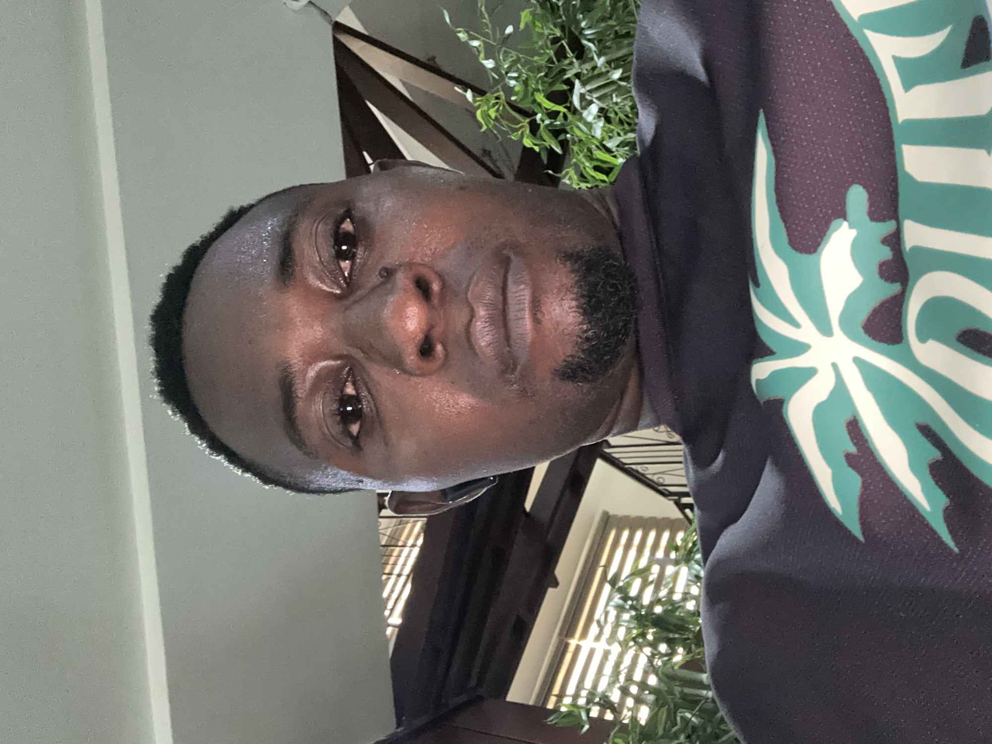 A dark-skinned man with short black hair and a goatee is shown in a close-up selfie. He is wearing a dark t-shirt with a white and teal graphic design on the front. He has an earbud in his left ear. The background appears to be indoors, with a ceiling visible at the top and some greenery and wooden structures in the distance. The lighting suggests it is daytime. The image does not provide any specific clues to suggest it was taken in Abuja, Nigeria.