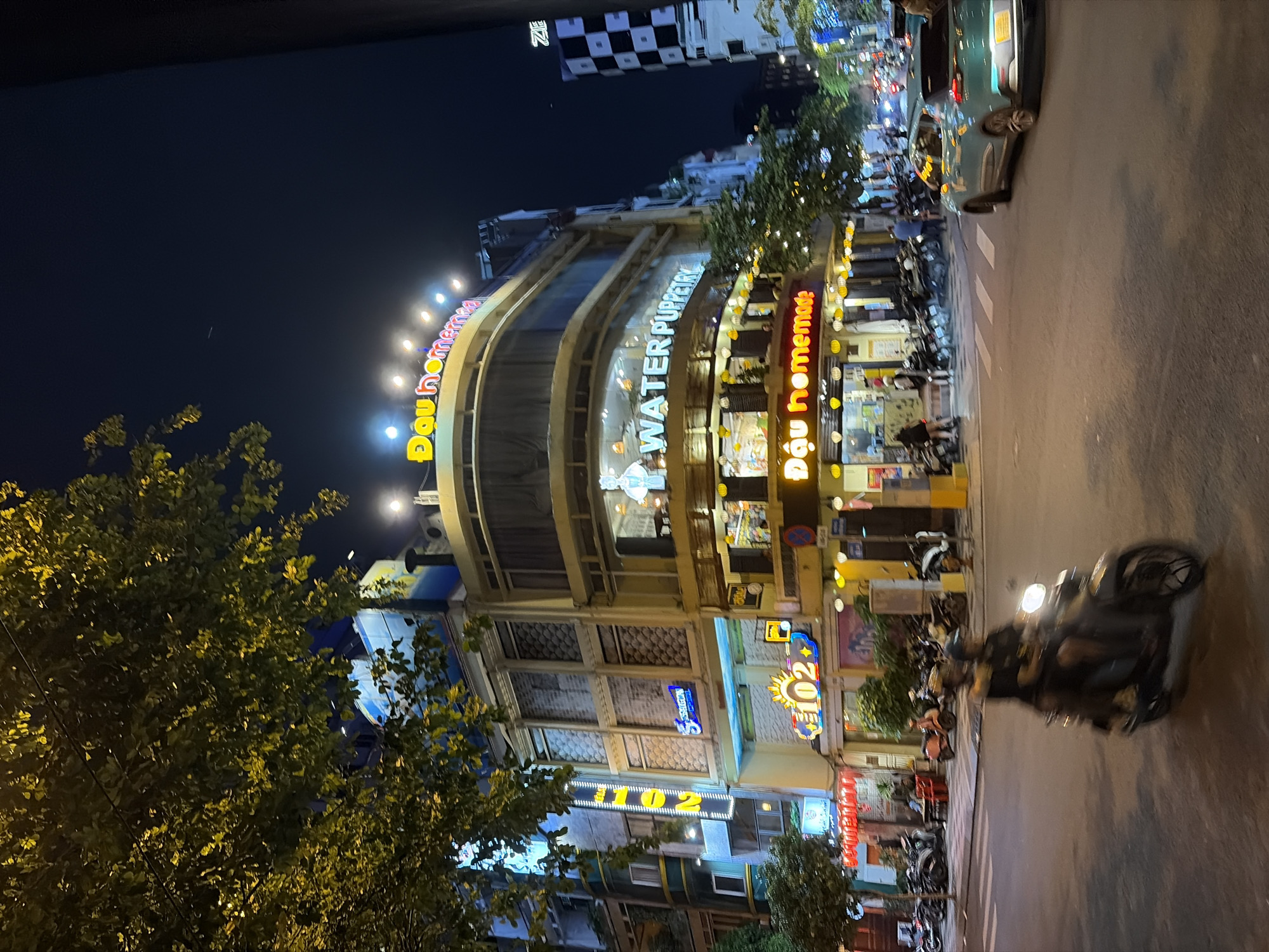 This image captures a nighttime scene on a street in Thủ Đức, Vietnam, featuring a multi-story building with illuminated signage. The building's facade is adorned with bright yellow and red neon signs, one prominently displaying "Đậu homemade" in yellow capital letters and another with "102" in neon blue and yellow. The building's lower levels appear to house businesses with windows showcasing interior lights and some customers. Above, a curved section of the building displays a sign reading "WATER PUPPETRY." A large tree with yellow-lit leaves extends into the frame from the left, partially obscuring the upper portion of the building. In the foreground, a motorcycle with two occupants is captured in motion on the road, with the rider and passenger both wearing helmets. Other parked motorcycles are visible on the sidewalk. A teal-colored car is also turning on the street. The overall environment suggests a busy urban setting at night, with an abundance of artificial lighting.