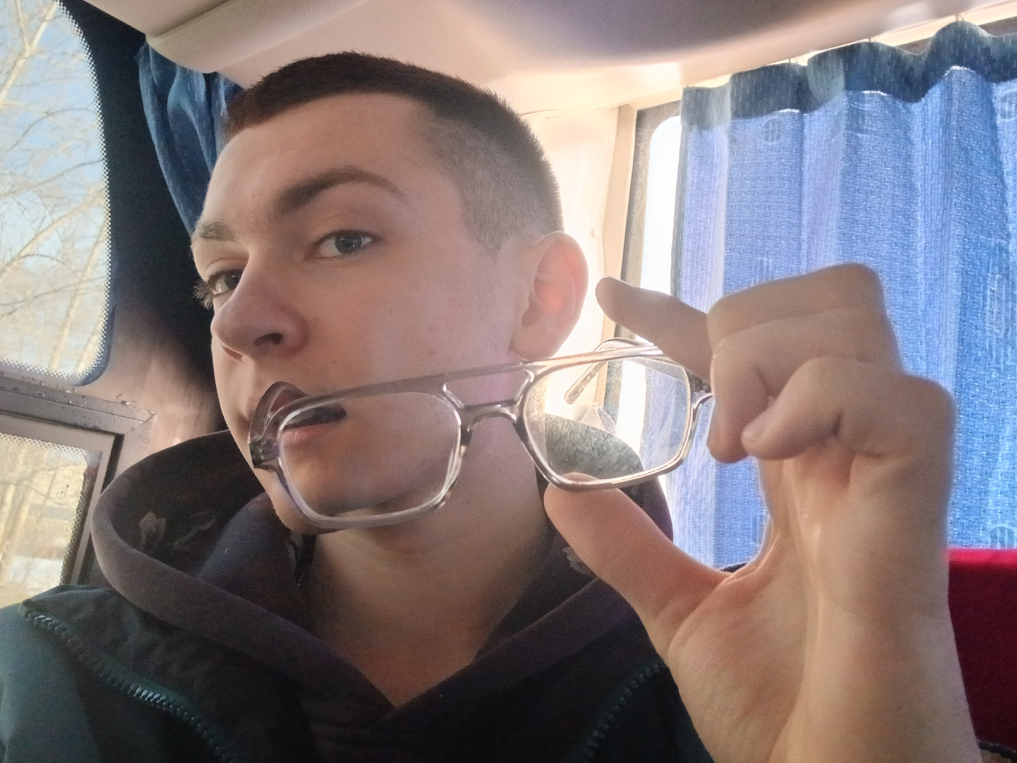A fair-skinned young man with short, dark hair is inside what appears to be a bus. He is holding a pair of glasses in his right hand and part of the frame is in his mouth. He's wearing a dark-colored hooded sweatshirt or jacket. Behind him are blue curtains, and a window is partially visible to the left. A window is behind the blue curtains as well.
There is no information about the location of the city of Berezniki, Russia to identify where the photo was taken.