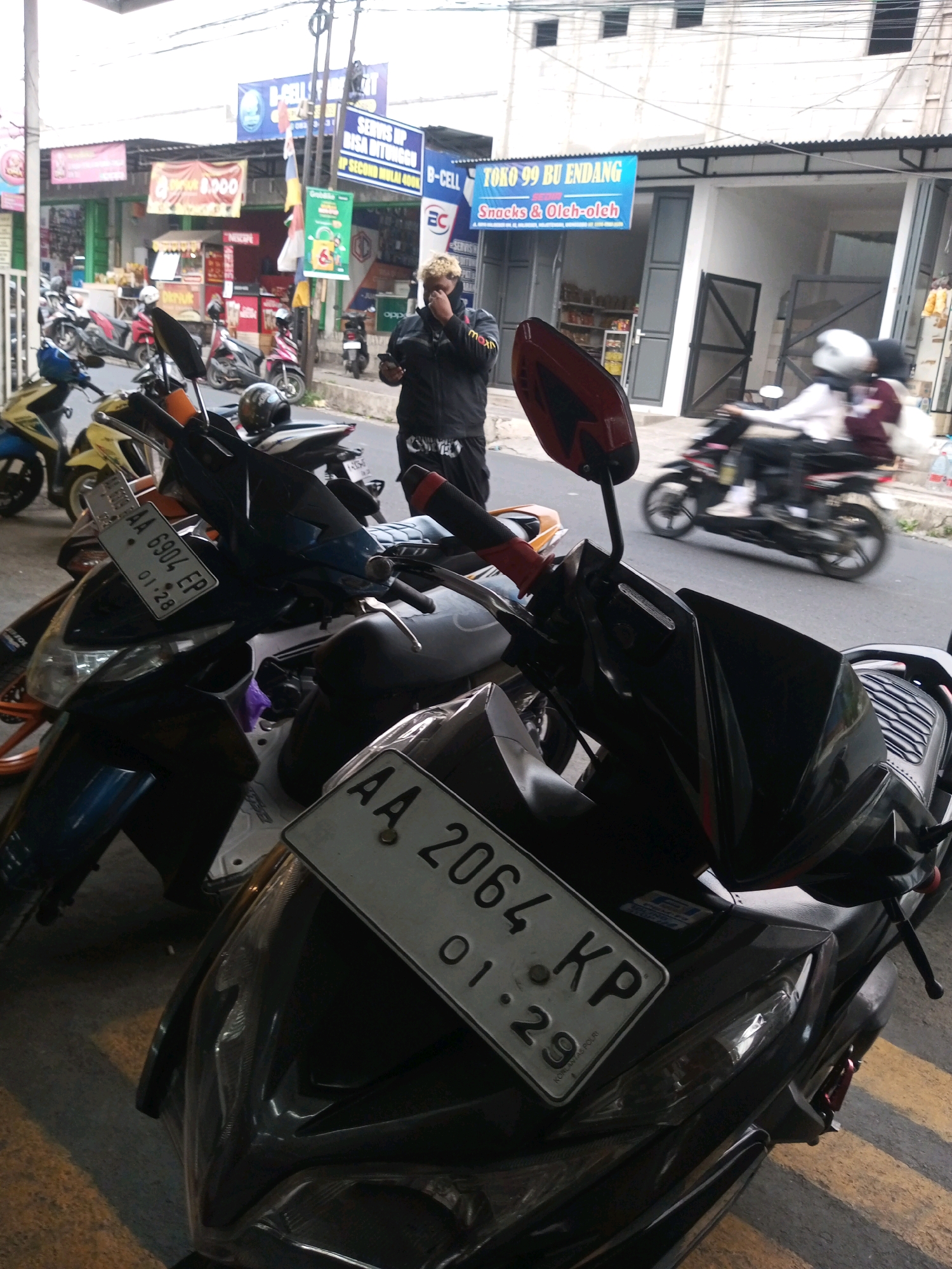 A man with bleached blonde hair, wearing a black jacket and pants, stands on a street in Mojotengah, Indonesia, looking at his phone. He is surrounded by parked motorcycles. In the background, a store with a blue sign reads "TOKO 99 BU ENDANG SNACKS & OLEH-OLEH". Another store to the left has a red banner with text that is difficult to read. Other motorcycles are visible on the street, with one in motion in the background. The scene appears to be a commercial area with shops and parking for vehicles.