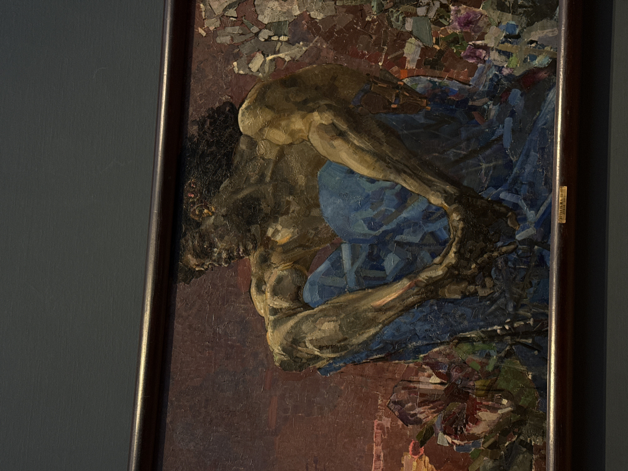 A framed painting depicting a nude man with dark hair. The man is contorted with his arms around his legs. He appears to be sitting or crouching. The man is rendered with heavy brushstrokes in shades of brown, tan, and blue, with a background of dark red and brown mixed with patches of green and blue. The painting appears to be housed in a museum. The location is likely Moscow, Russia, as suggested by the Cyrillic text on the frame.