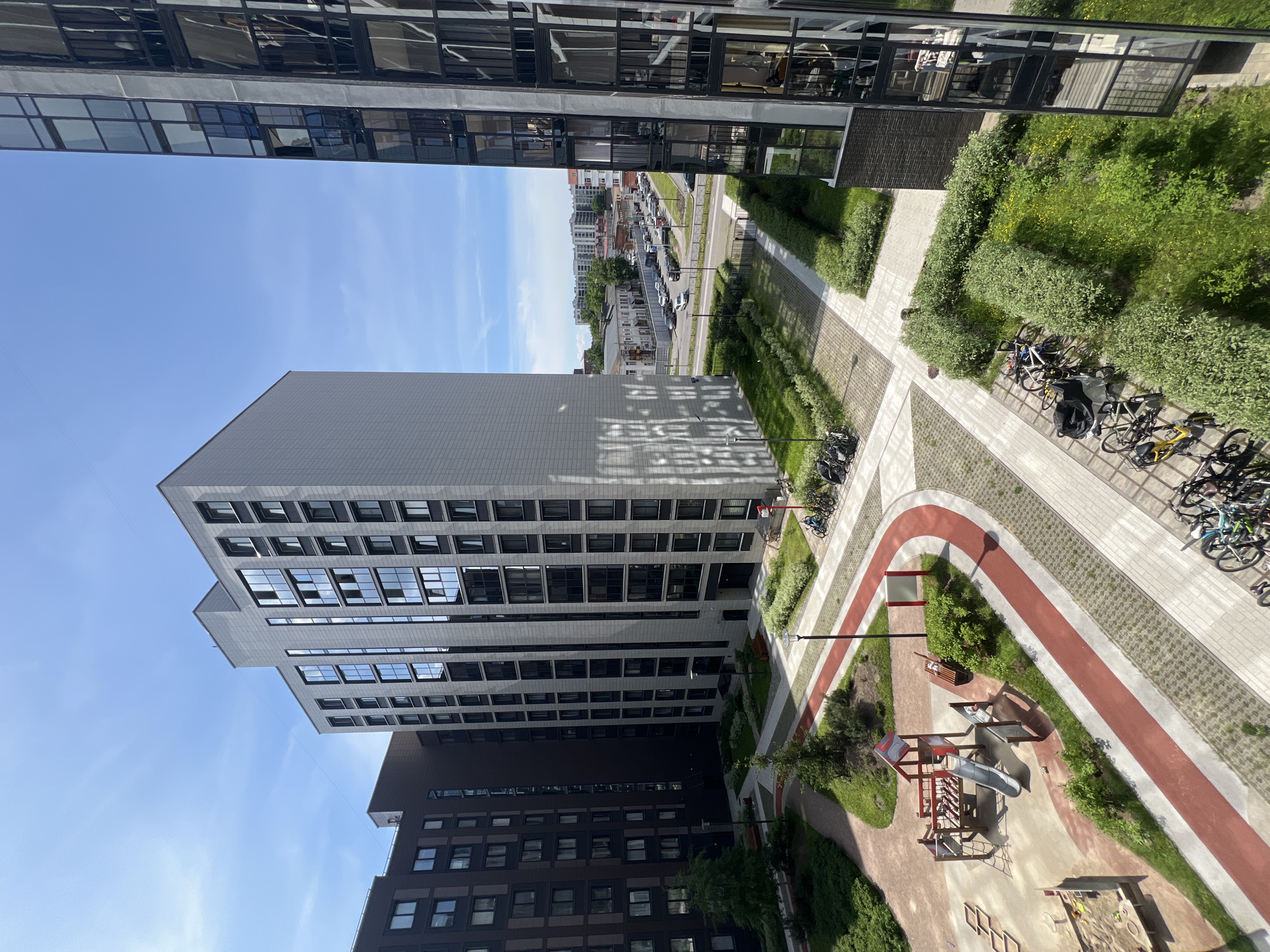 This is a high-angle, medium shot photograph of a modern residential courtyard in Saint Petersburg, Russia, on a clear, sunny day. The courtyard is bordered by several multi-story apartment buildings, featuring contemporary architecture with light-colored facades and large windows. In the foreground, to the left, is a children's playground with a slide, swings, and a sandpit. Adjacent to the playground is a paved pathway with red and white markings, winding through landscaped areas with green grass and bushes. Several bicycles are parked in various locations around the courtyard. In the background, a street with traffic can be seen, with more buildings and trees lining the road. The sky is a vibrant blue with a few wispy clouds. The overall scene depicts a well-maintained urban residential area with amenities for recreation.