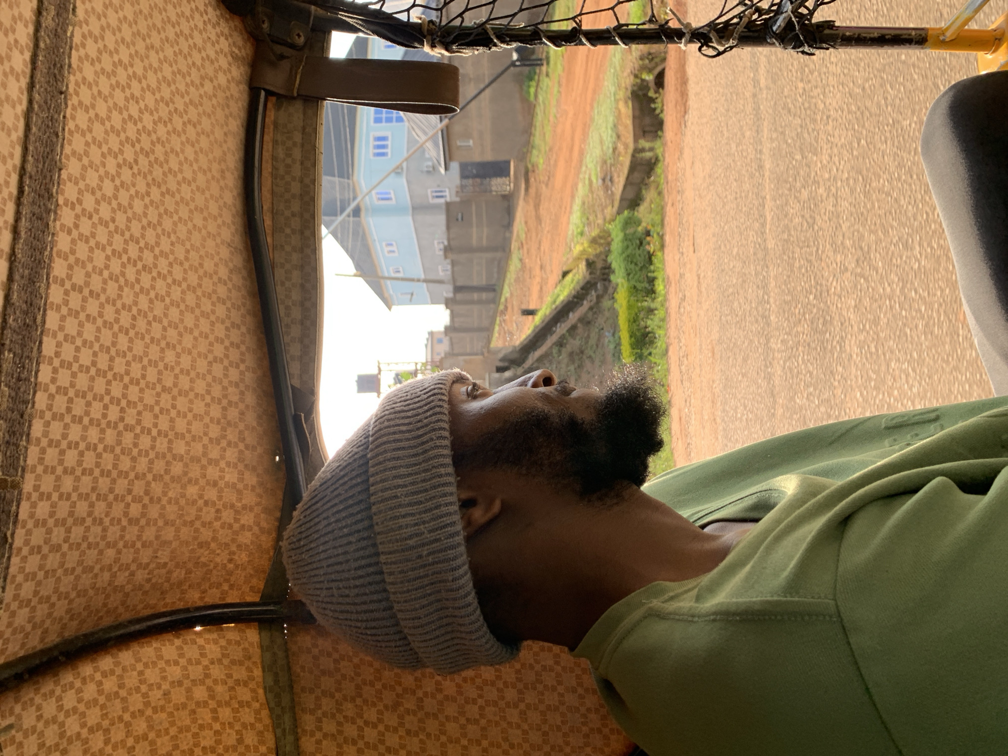 A man wearing a grey beanie and a green long-sleeved shirt is sitting in the back of a tuk-tuk, looking out the side. The interior of the tuk-tuk has a textured, patterned beige ceiling. Outside, a dusty road is visible, with buildings and greenery in the background. The man has a beard and is positioned in profile. The image appears to be taken in Gura Top, Nigeria, given the architectural style and the presence of a tuk-tuk.
