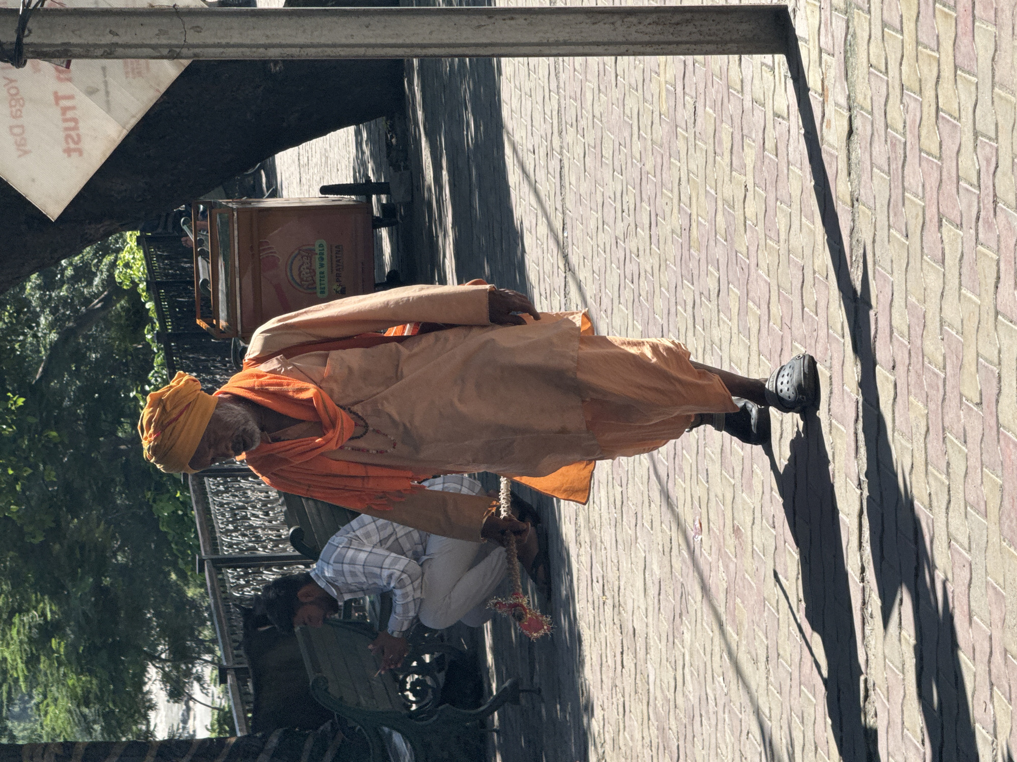 In Rishikesh, India, a man in an orange turban and robes walks along a paved walkway. He is holding a red and pink tasseled object by a string in his right hand. In the background, another man in a checkered shirt and grey pants is sitting on a bench and looking at his phone. A metal cart with the words "BETTER WORLD" and "PRAYATNA" is visible behind the man in orange robes. The scene is illuminated by sunlight, casting shadows on the walkway.