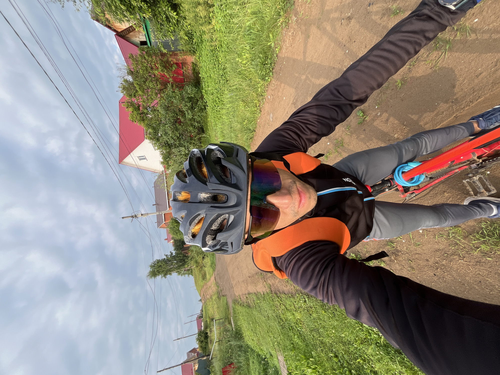 A man in a black cycling helmet, reflective sunglasses, and a black long-sleeved jersey with orange shoulder pads is riding a red bicycle on a dirt road in the city of Chaikovsky, Russia. The road curves uphill, bordered by green grass and vegetation. Several houses with red roofs are visible in the background, along with utility poles and power lines. The sky is cloudy. The image appears to be a selfie taken by the cyclist.