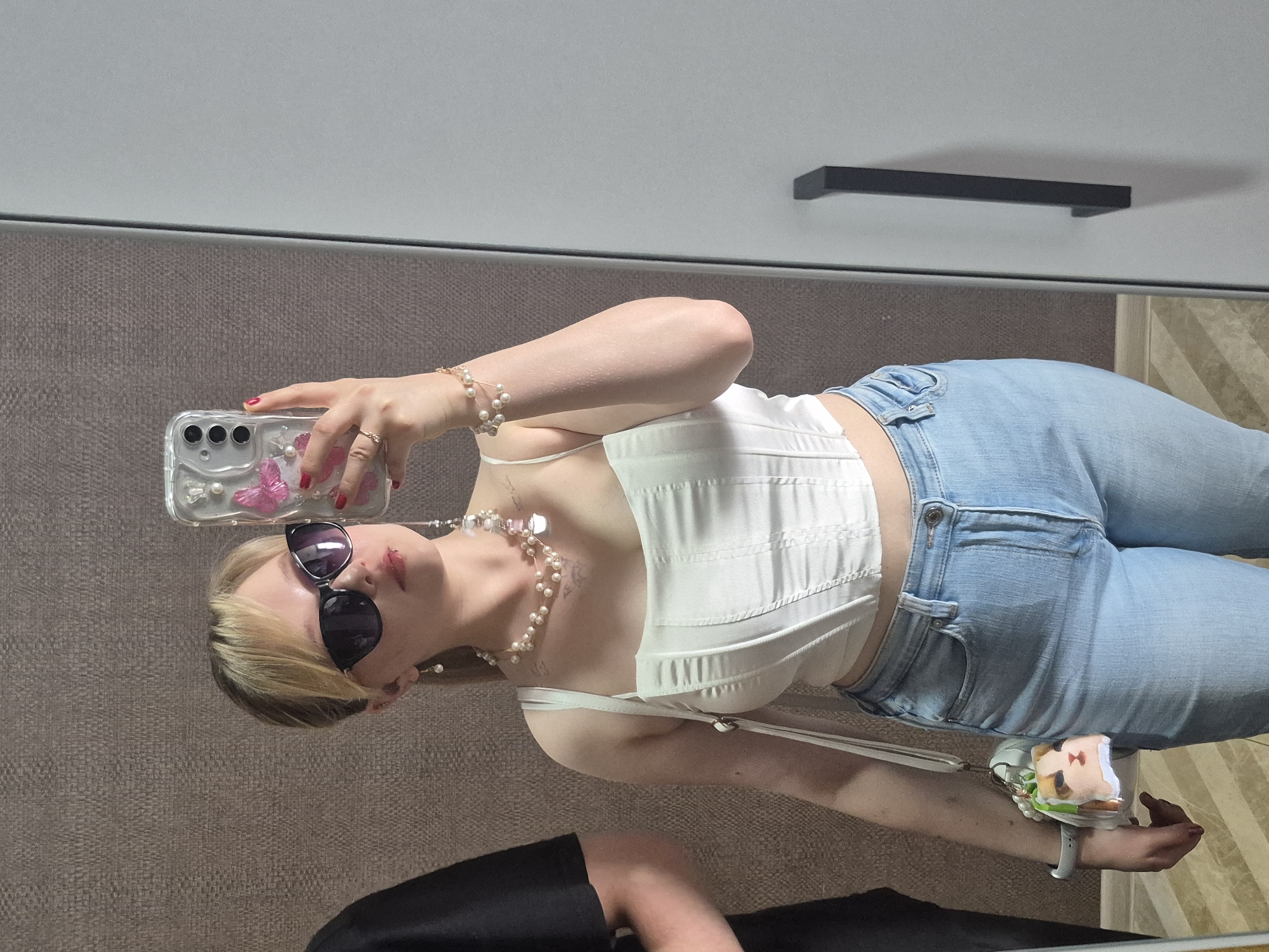 A young Caucasian woman with blonde hair is taking a selfie in what appears to be a fitting room or a large mirror. She is wearing a white corset top, light-wash denim jeans, large black sunglasses, and multiple pearl accessories including earrings, a necklace, and a bracelet. A phone with a pink butterfly design is held in her right hand. A small white purse with a cat-shaped charm is slung over her left shoulder. The background consists of a textured brown wall and a grey cabinet with a black handle. The lighting is bright, suggesting it might be daytime. There is no visible indication of the city of Izhevsk, Russia.