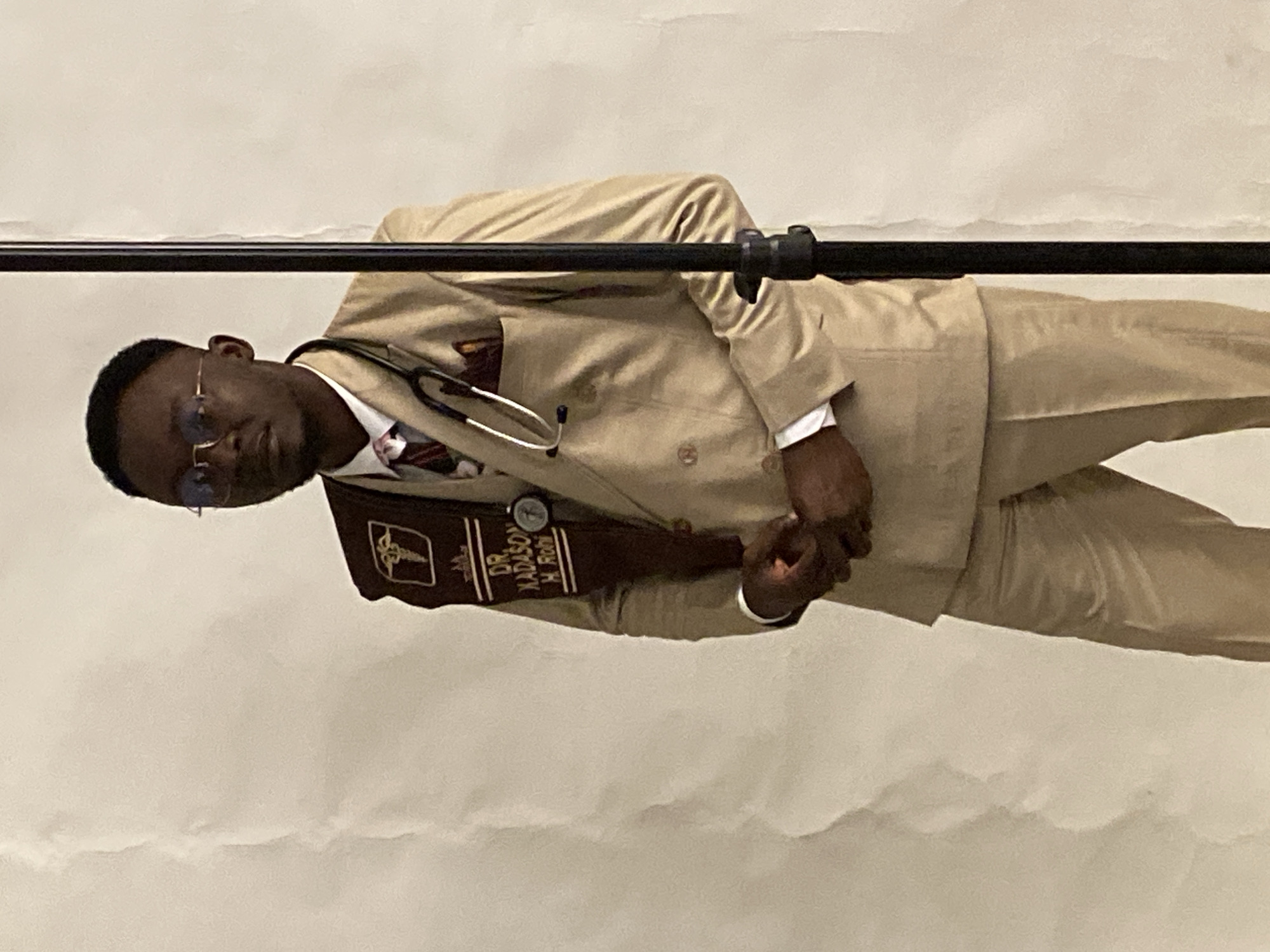 A full-shot, medium-skinned Black man, appearing to be in his late 20s or early 30s, stands facing the camera. He wears a tan suit with a double-breasted jacket, a white dress shirt, and a patterned tie. A stethoscope is draped around his neck, with its earpieces resting on his collar and the diaphragm tucked into his chest pocket. He also wears round-rimmed glasses. A dark brown sash is draped over his left shoulder and across his chest, featuring a caduceus medical symbol and embroidered text that reads "DR. XADASON H. ROHI." His hands are clasped in front of him. He stands against a white, textured backdrop, with a thin black pole positioned to his right. The lighting is even, with some minor shadows cast on the backdrop. The image appears to have been taken indoors, possibly in a studio setting in Jos, Nigeria.