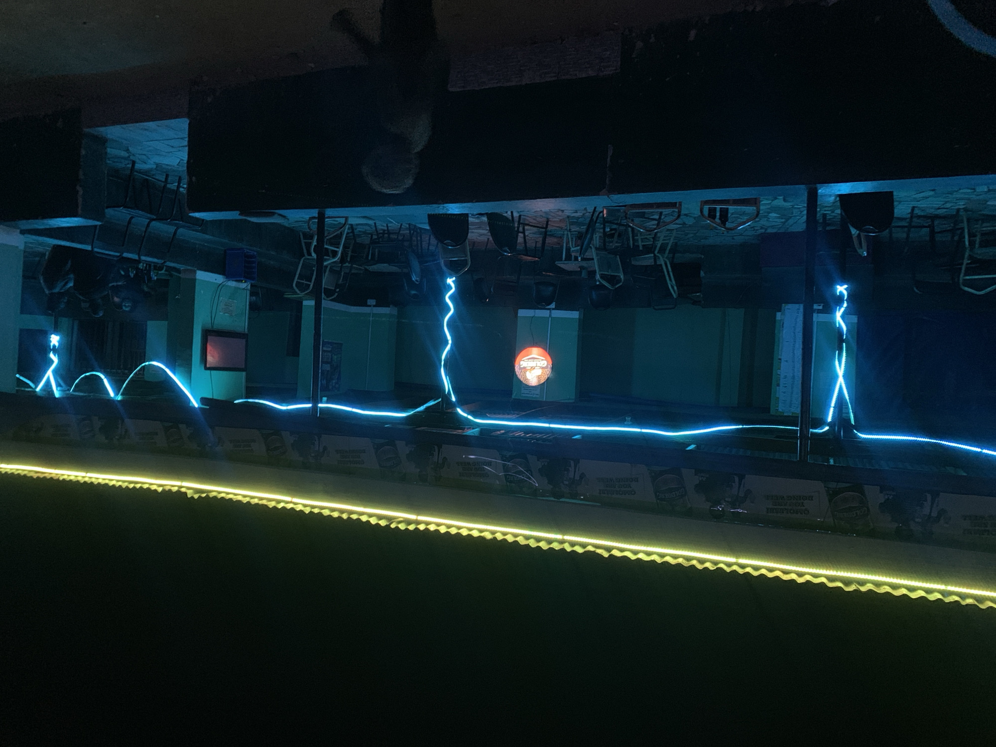 The image depicts an interior scene, possibly a bar or restaurant, located in New Stadium, Nigeria, taken upside down. The space contains rows of chairs and tables arranged in a dining setup. Above, the ceiling is dark and nondescript, with some hanging objects that appear to be chairs stacked upside down. Below, there is a stage area lit with blue neon lights in a wavy pattern, and a strip of yellow lights at the bottom edge. A sign with the word "COLDERS" is visible on the wall facing the camera.