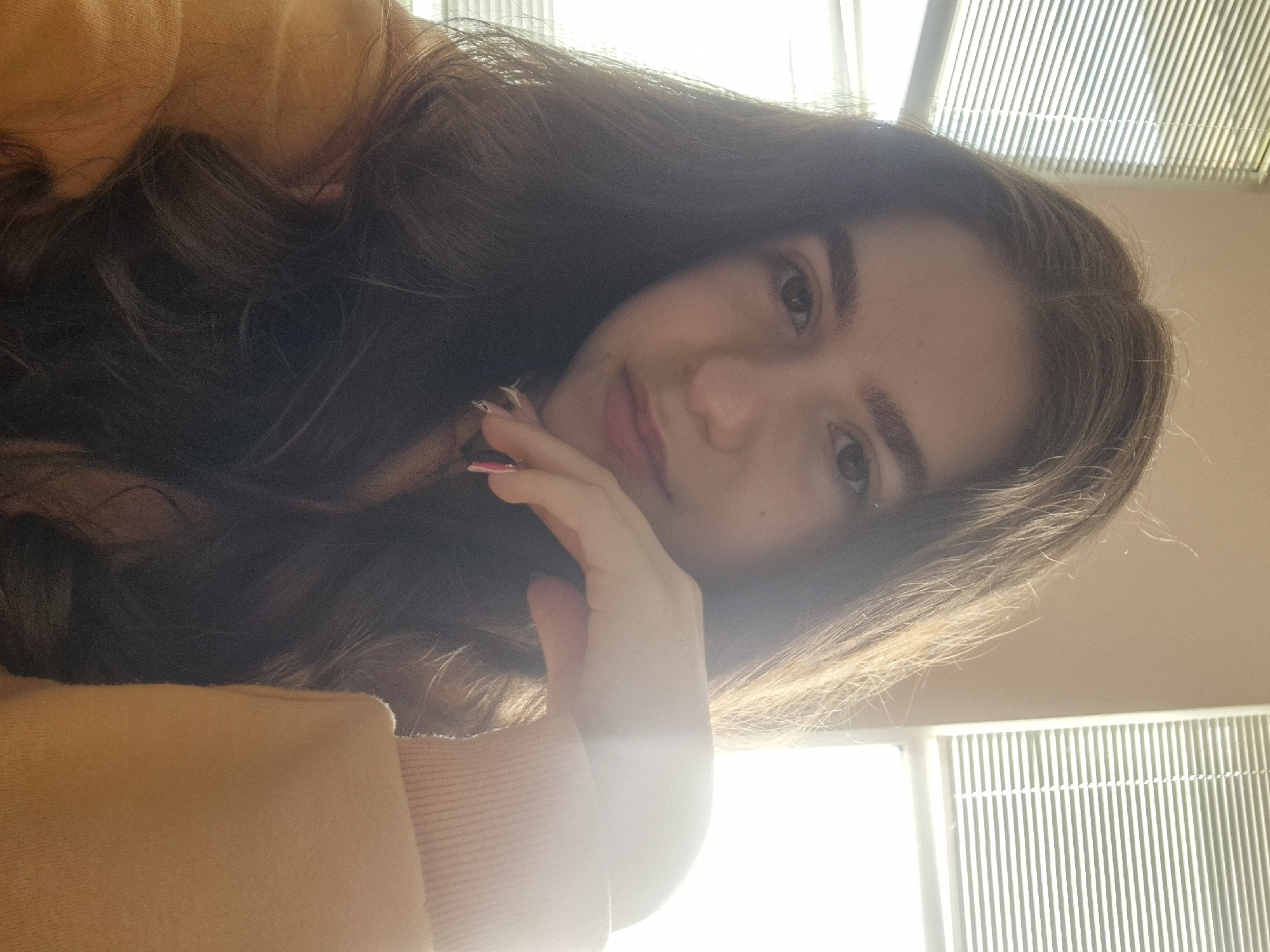 A young woman with long, wavy brown hair poses for a selfie in a brightly lit room. She rests her chin on her right hand, displaying nails with a patterned design. She is wearing a mustard-colored sweater. Behind her, a window with horizontal blinds is visible. Location in Pavlikeni, Bulgaria cannot be determined based on the image.