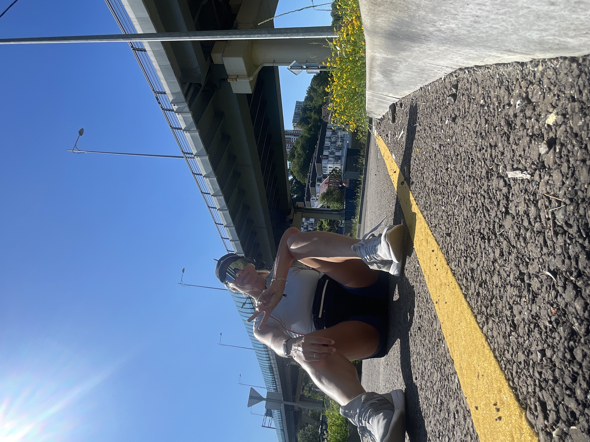 An individual is captured sitting on an asphalt path under a large concrete overpass or bridge in Sochi, Russia. The person, dressed in a white top, dark shorts, and white athletic shoes, is wearing large mirrored sunglasses and possibly a visor or cap. They are seated with their knees bent, making a peace sign (V-sign) with one hand and looking towards the camera.
The scene is set outdoors on a bright, sunny day, evidenced by the clear blue sky and a prominent sun flare in the lower left of the image. The path beneath the overpass features a distinct yellow dividing line. To the right, a patch of yellow flowers blooms next to a concrete barrier. In the background, beyond the overpass structure, green trees and urban buildings are visible, suggesting a city environment. A street lamp is also present under the bridge. The individual's pose and gesture convey a relaxed and possibly cheerful mood. No visible text is present in the image.