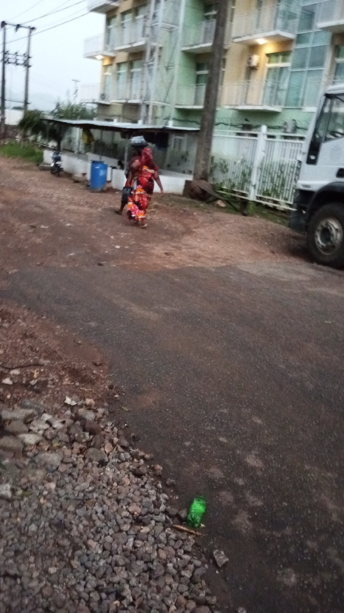 The photograph depicts a street scene in Adankolo, Nigeria. An African woman in a colorful red dress carries an item on her head. She walks beside another person down a partly unpaved road. A building is behind her and includes green paint and white balconies. A white truck is partially visible on the right side of the frame. Debris and a green bottle are on the side of the road in the lower portion of the image.