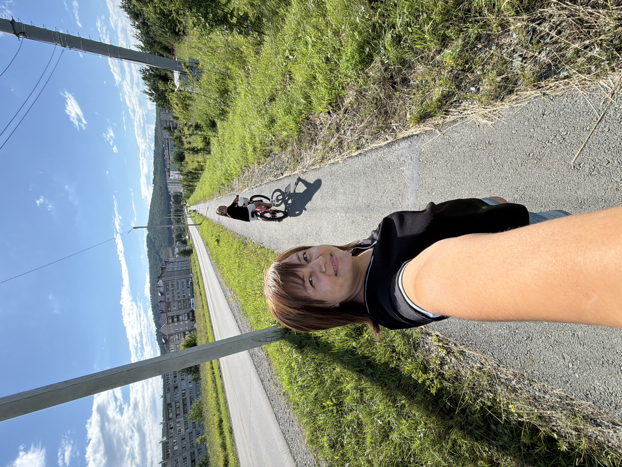 A woman and a young girl are walking on a paved path next to a road in a residential area. The woman is in the foreground, holding her phone up to take a selfie. She has brown hair and is wearing a black shirt and denim shorts. The girl is in the background, riding a pink bicycle. She has dark hair and is wearing a black shirt and white shorts. The road is lined with grass and trees, and in the distance, there are several apartment buildings and a wooded hillside. The sky is blue with white clouds, and the sun is shining.