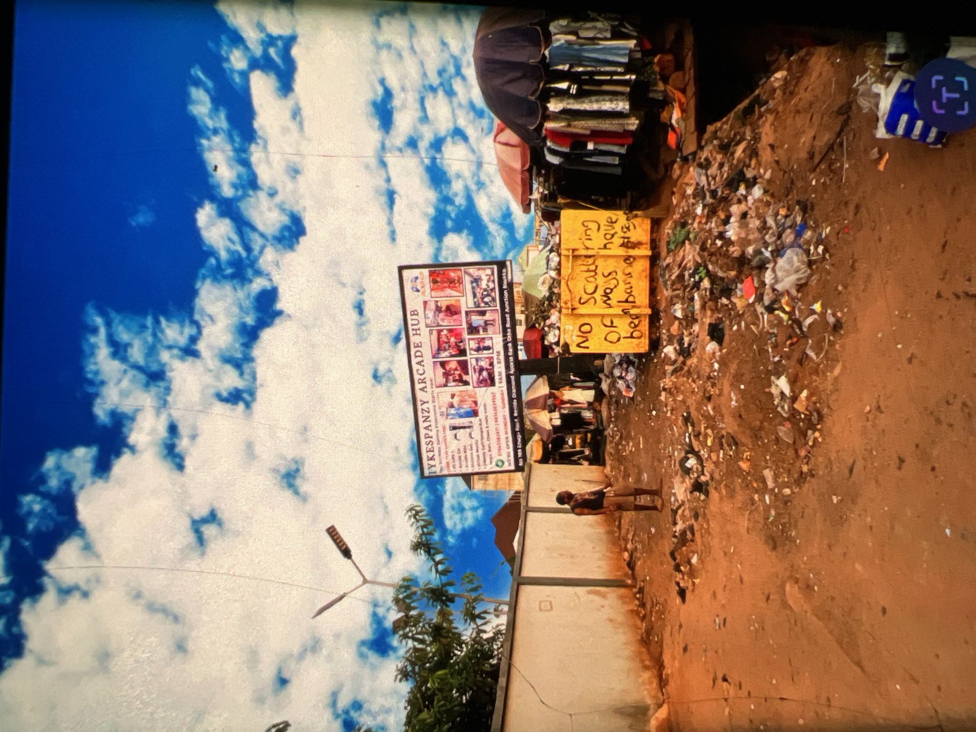 The image depicts a street scene in Lagos, Nigeria. In the foreground, a dirt path is littered with an extensive amount of garbage, creating a mound of refuse. A single man stands in the middle of this path, looking towards the right side of the frame. Further back, a white wall runs along the left side of the scene, with a street light visible above it. A large billboard is mounted on a structure behind the wall, advertising "IYKESPANZY ARCADE HUB" and showcasing various products. To the right of the billboard, market stalls are set up with umbrellas providing shade, displaying clothing items for sale. A yellow dumpster with handwritten text that reads "NO Scattering OF ways have been banned" is also visible amongst the trash. The sky above is a vibrant blue, dotted with white clouds. The overall impression is one of a bustling, albeit untidy, commercial area within the city.