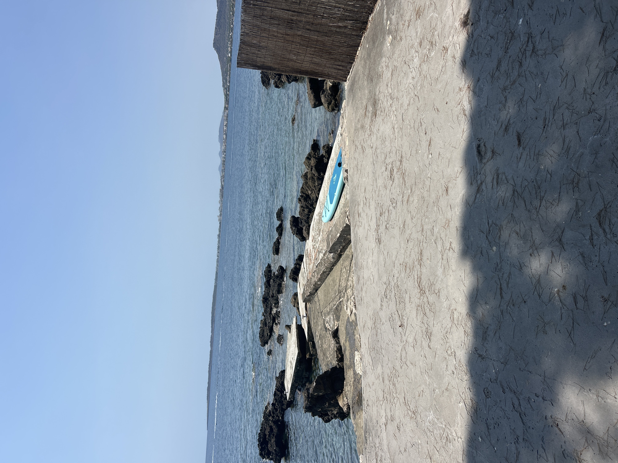 The image depicts a concrete pier extending into the clear blue sea under a bright, cloudless sky. The pier is partially submerged and weathered, with rocks jutting out of the water around it. A blue paddleboard rests on the concrete surface of the pier, positioned to the right of the frame. To the right of the paddleboard, a tall, dark bamboo fence partially obstructs the view of the sea. In the background, a distant coastline with buildings and hills is visible across the water, suggesting the location is near a populated area. The foreground of the image shows the concrete ground of the pier, with fallen pine needles scattered across its surface and a shadow cast from an unseen object, likely foliage. The image captures a serene, sunlit scene in what appears to be a coastal resort area.