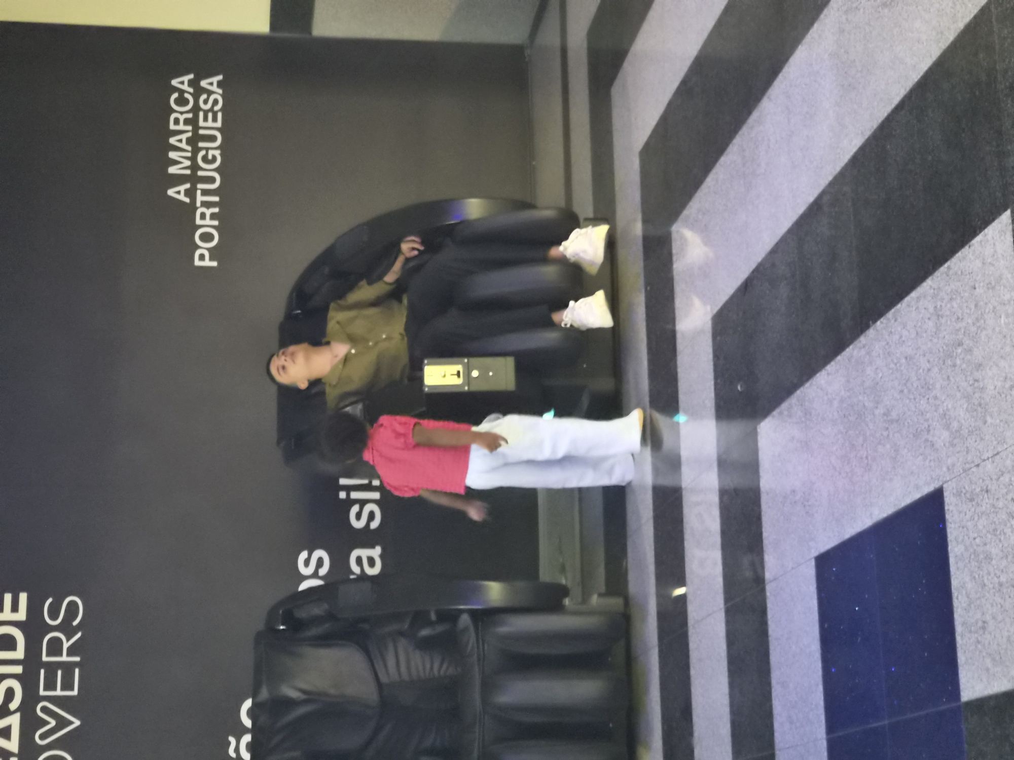 In Bragança, Portugal, two individuals are present within a public space, possibly a commercial establishment or waiting area. In the foreground, a young child, with dark curly hair, stands facing away from the camera. The child is wearing a short-sleeved pink top and white trousers. To the child's right, a person, who appears to be an adult male with short dark hair, is seated in a large, dark-colored reclining massage chair. The man is wearing a green collared shirt and dark trousers, with white sneakers on his feet. His eyes are closed, suggesting he may be resting or experiencing the massage. A control panel is visible on the armrest of the chair. To the left of the scene, a portion of another identical dark-colored massage chair is visible. The background features a black wall with white lettering. The floor is tiled with a pattern of alternating black and grey stripes, with a dark blue square section in the lower left corner. The lighting appears to be artificial, contributing to the indoor atmosphere of the space.
