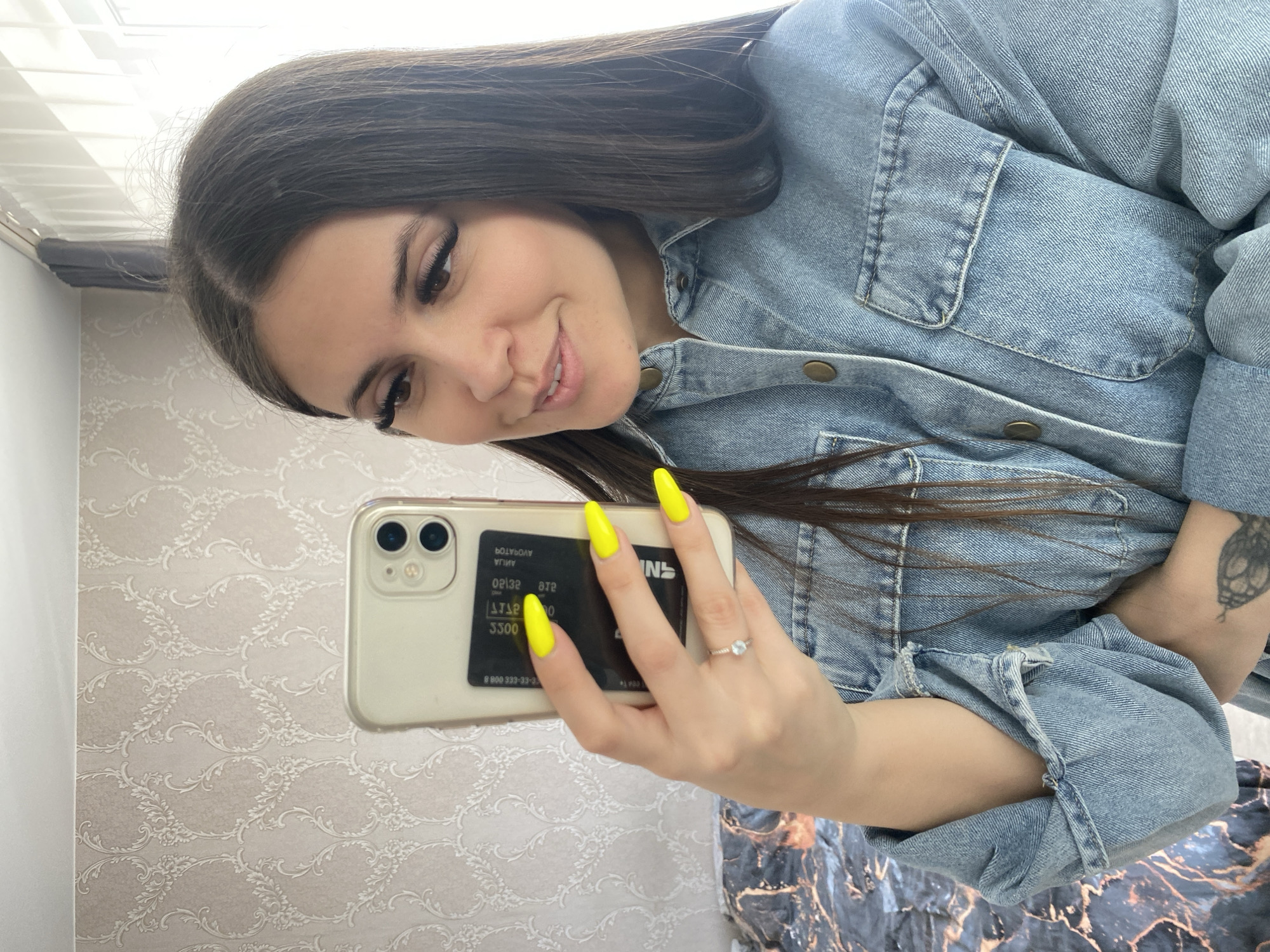 A woman is taking a selfie indoors. She has long, dark brown hair and is wearing a denim jacket. She is holding a cell phone with bright yellow nails. A credit card is placed on the back of the phone. Her left arm has a tattoo. The background consists of a white wall with a patterned wallpaper and a glimpse of a marble-patterned surface. The location is likely indoors, but the specific location in the city of Chaikovsky, Russia, cannot be determined from the given image.
