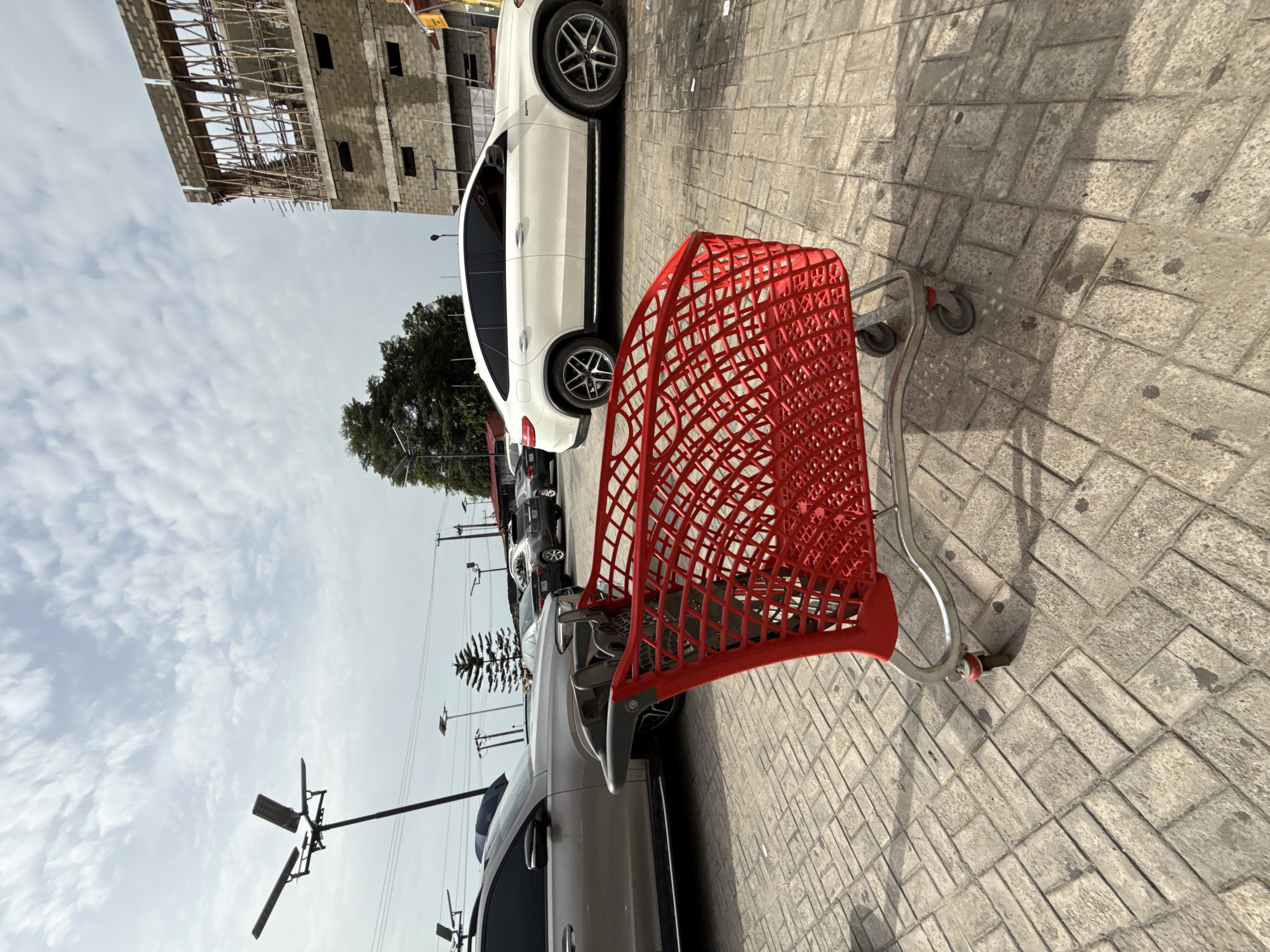 A shopping cart is positioned on a stone-paved surface in Ado, Nigeria, in a parking lot adjacent to several cars and a building under construction. The cart is predominantly red, with a silver handle and wheels. The cars in the parking lot include a white SUV and other various models in darker shades. The building under construction is visible in the background. Overhead, a cloudy sky is seen with utility poles and lines lining the street.