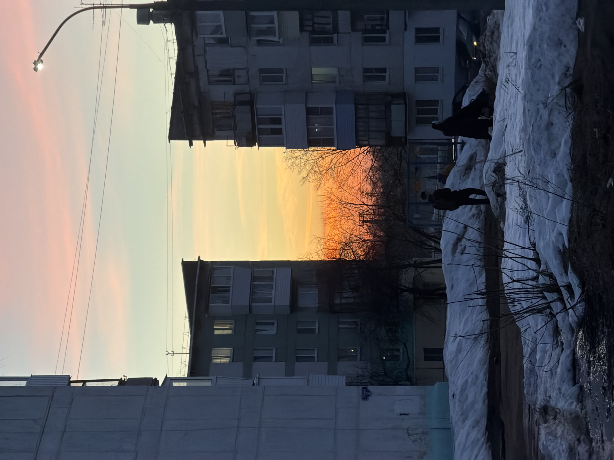 The image depicts an urban scene in Chaikovsky, Russia, at dusk during winter. Two multi-story apartment buildings stand on either side of a street or open area. The sky between the buildings is a gradient of orange and yellow hues, suggesting a sunset. The foreground shows a snow-covered area, with some bare branches and vegetation visible through the snow. Two dark figures, possibly people, are situated on the snow bank. A street lamp stands in the upper left corner of the image, with power lines connecting it to the buildings. The buildings are typical Soviet-era concrete structures with rows of windows.