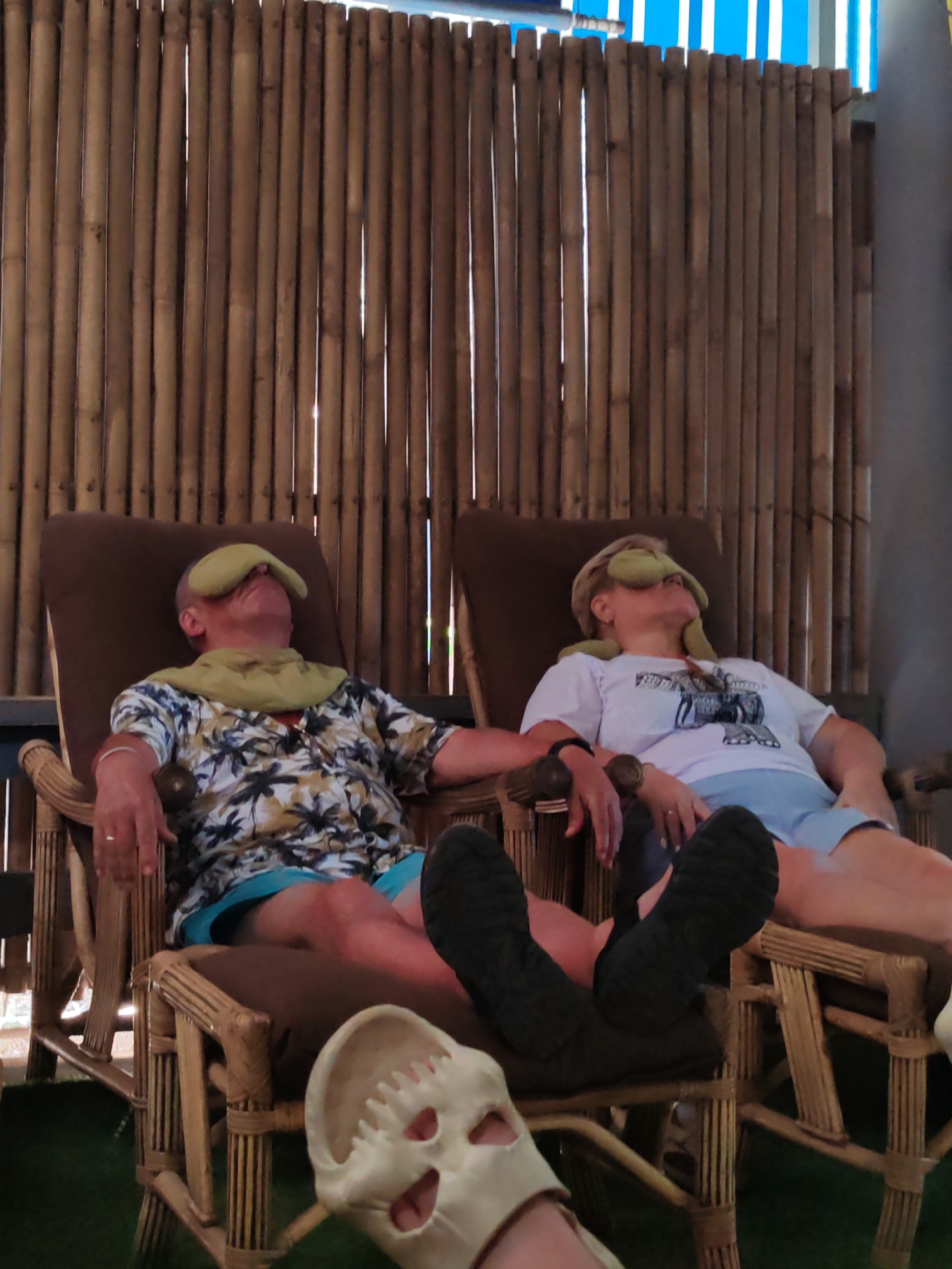 In Ban Khlong Khian, Thailand, two individuals are relaxing in what appears to be an outdoor spa or relaxation area. They are seated in wicker-style chairs with brown cushions, positioned in front of a tall, bamboo-like screen or wall. Both individuals have green eye masks and neck pillows. The person on the left is wearing a short-sleeved shirt with a tropical print, while the person on the right wears a white t-shirt with a graphic print. Both are wearing blue shorts. Their feet are positioned forward in the chairs. A sandal, possibly a Croc, is in the lower foreground. The chairs sit on an artificial green surface or grass. The overall scene suggests a tranquil, tourist-oriented setting.