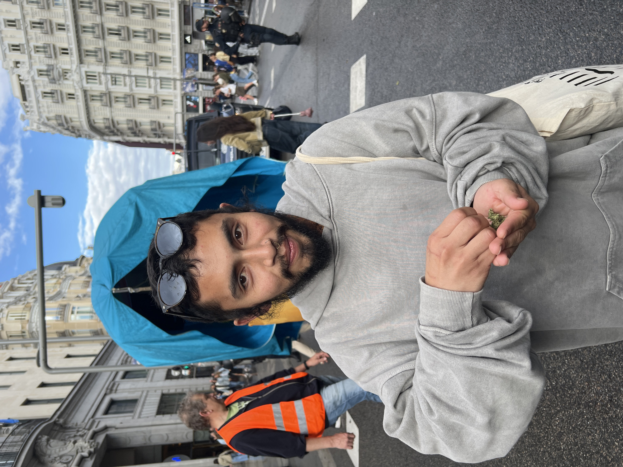 A medium close-up shot shows a person with a beard, a nose ring, and sunglasses perched on their head, wearing a gray hoodie, holding a small amount of green material in their hands. The person is standing on a street in Madrid, Spain. Behind them is a blue and yellow object, resembling a flag or banner. In the background, pedestrians walk along a crosswalk. Several police officers in tactical gear are present amidst the crowd. Buildings with classical architectural details are visible in the background, confirming the urban setting. An individual wearing a high-visibility orange vest is also walking nearby.