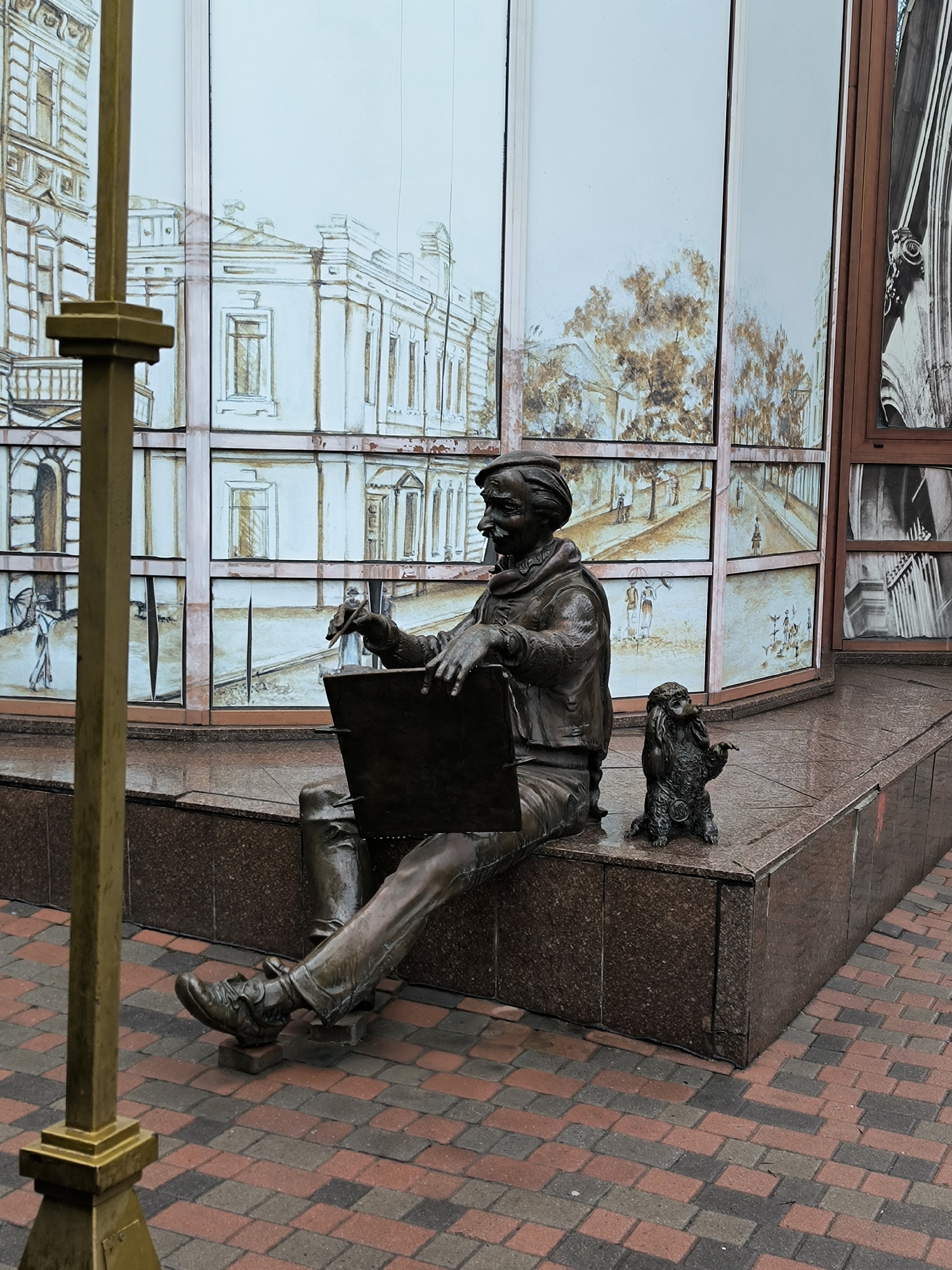 A bronze statue of a seated artist with a pencil in his right hand sits on a granite bench. The artist wears a cap and jacket. A small statue of a dog is on the bench next to the artist. A gold lamp post is positioned to the left. Behind the statue are large windows with images drawn on them that represent buildings and people. The ground is paved with red and gray bricks. This artwork is located in Vinnytsia, Ukraine.