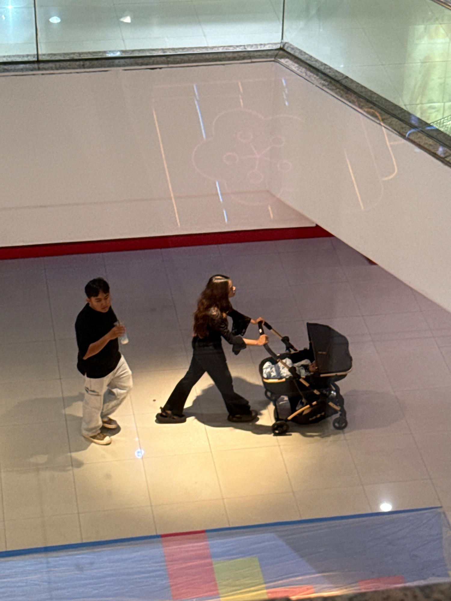 In a well-lit indoor environment, possibly a shopping mall with tiled flooring, a man and a woman are depicted. The man, wearing a black t-shirt and light-colored pants, walks to the left of the frame, holding a clear plastic bottle. To his right, the woman, dressed in black flared pants and a black top with bell sleeves, pushes a dark-colored baby stroller. The stroller appears to have a baby inside, partially visible. The background consists of a white wall with subtle reflective patterns and a railing at the top. The overall scene suggests a casual outing. This image does not contain any specific landmarks or clues that would pinpoint its location to Surabaya, Indonesia.