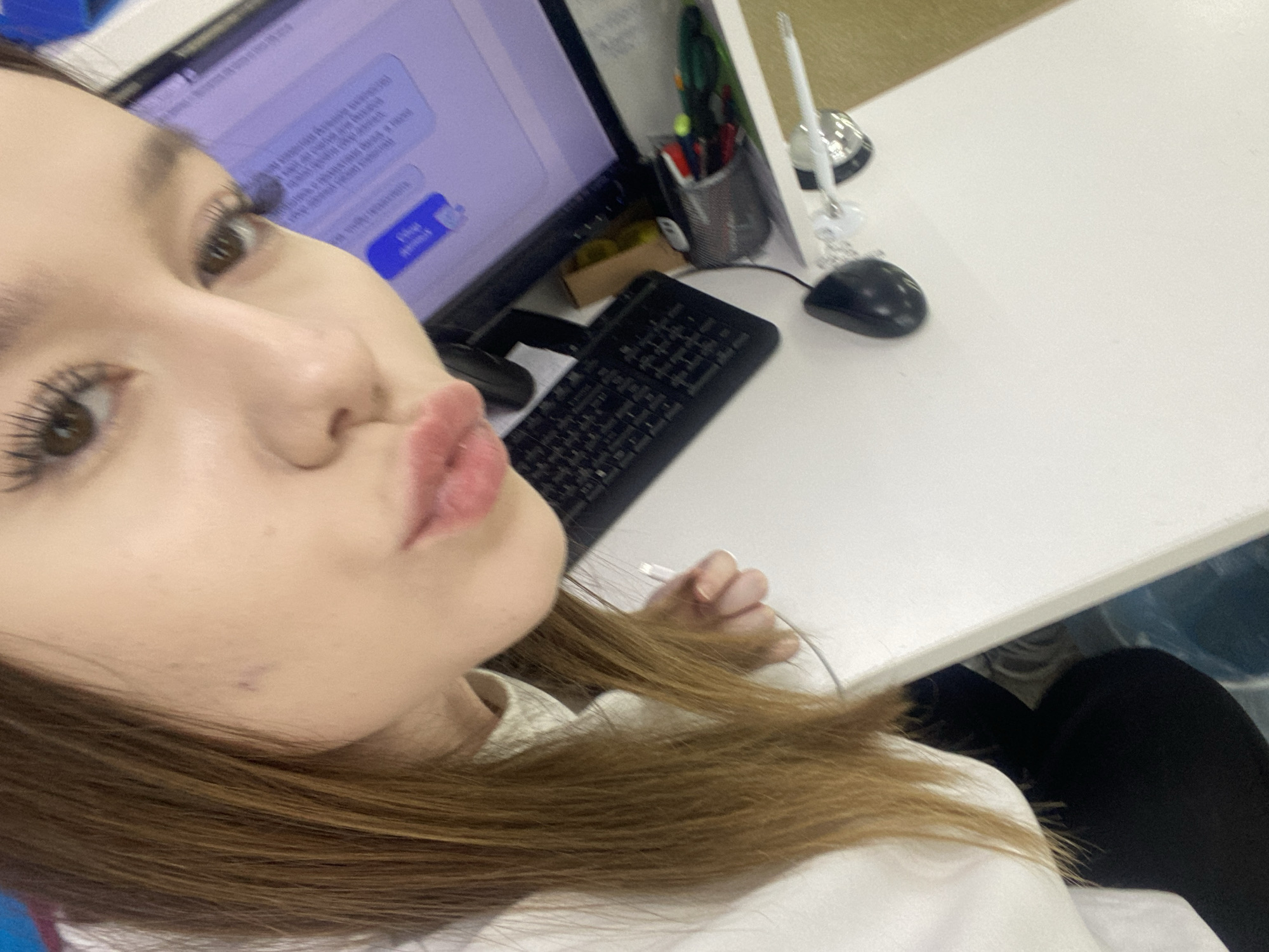 A young woman is taking a selfie at a desk. She is pouting her lips at the camera. She has brown hair and is wearing a white shirt. In the background, there is a computer monitor, a keyboard, a mouse, a pen holder with pens and pencils, and a white desk. The computer monitor displays a chat window. The location may be in an office or workplace in the city of Chaikovsky, Russia.